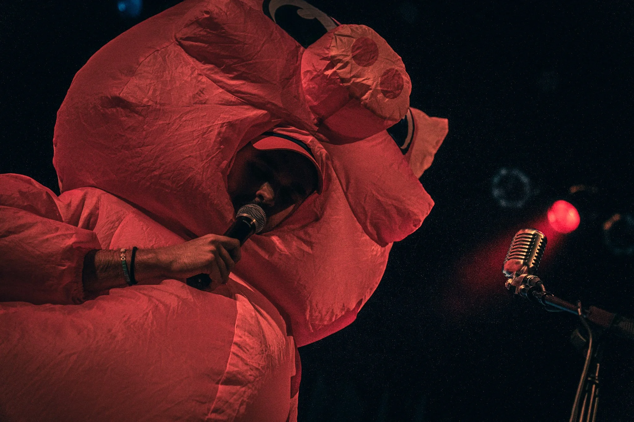 A person wearing a red hooded garment speaking into a microphone in a dark setting, with a vintage microphone visible in the foreground, surrounded by a starry sky background.