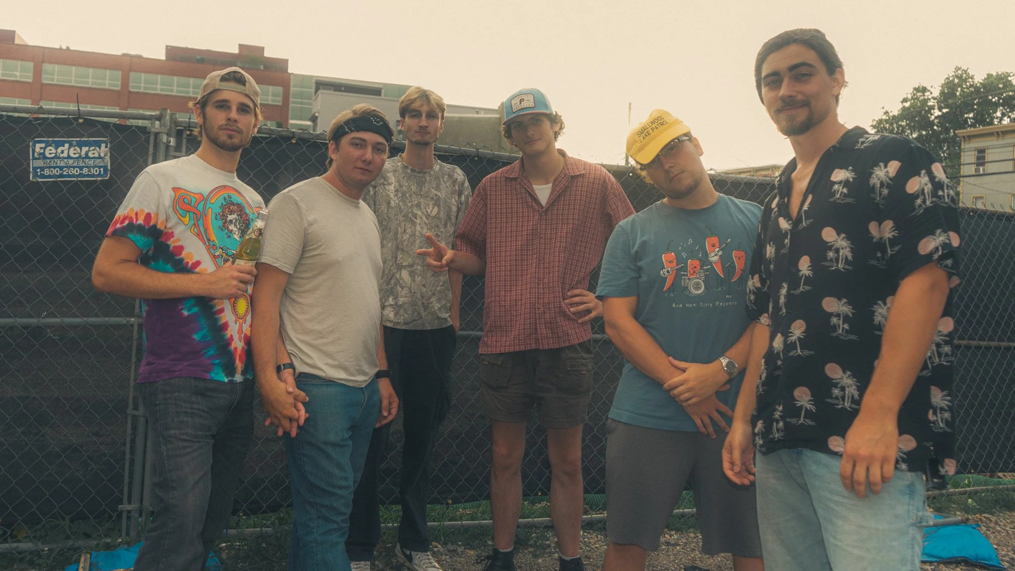 Six young men standing outdoors in front of a chain-link fence. They are dressed casually, some making peace signs or relaxed poses. Buildings are visible in the background.