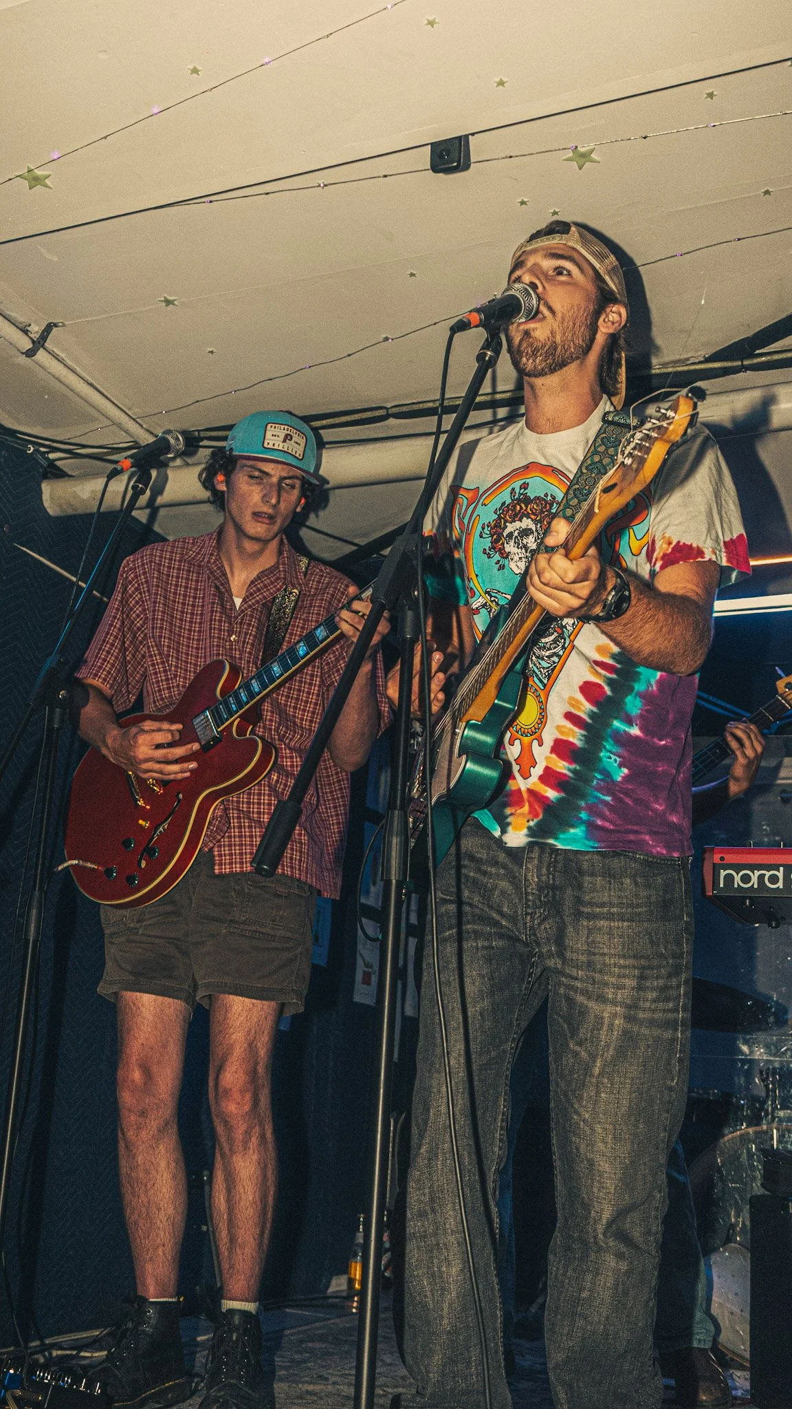 Two musicians performing on stage. The musician in the foreground is playing an electric guitar and singing into a microphone, wearing a colorful tie-dye T-shirt and gray plaid pants. The musician in the background is playing a red electric guitar, w
