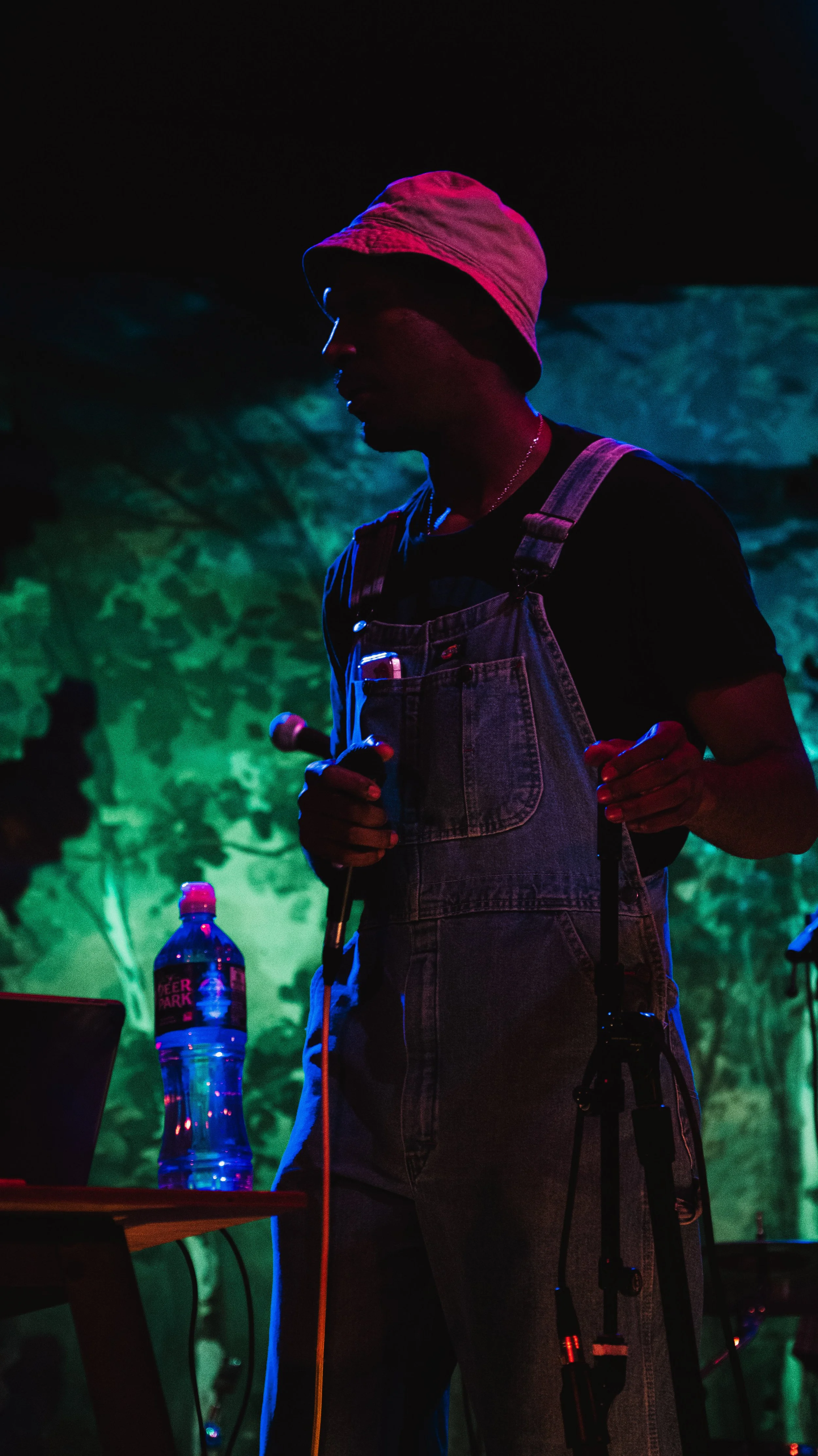 Person wearing a pink bucket hat and denim overalls holding a microphone in a darkly lit environment with colorful background lighting.