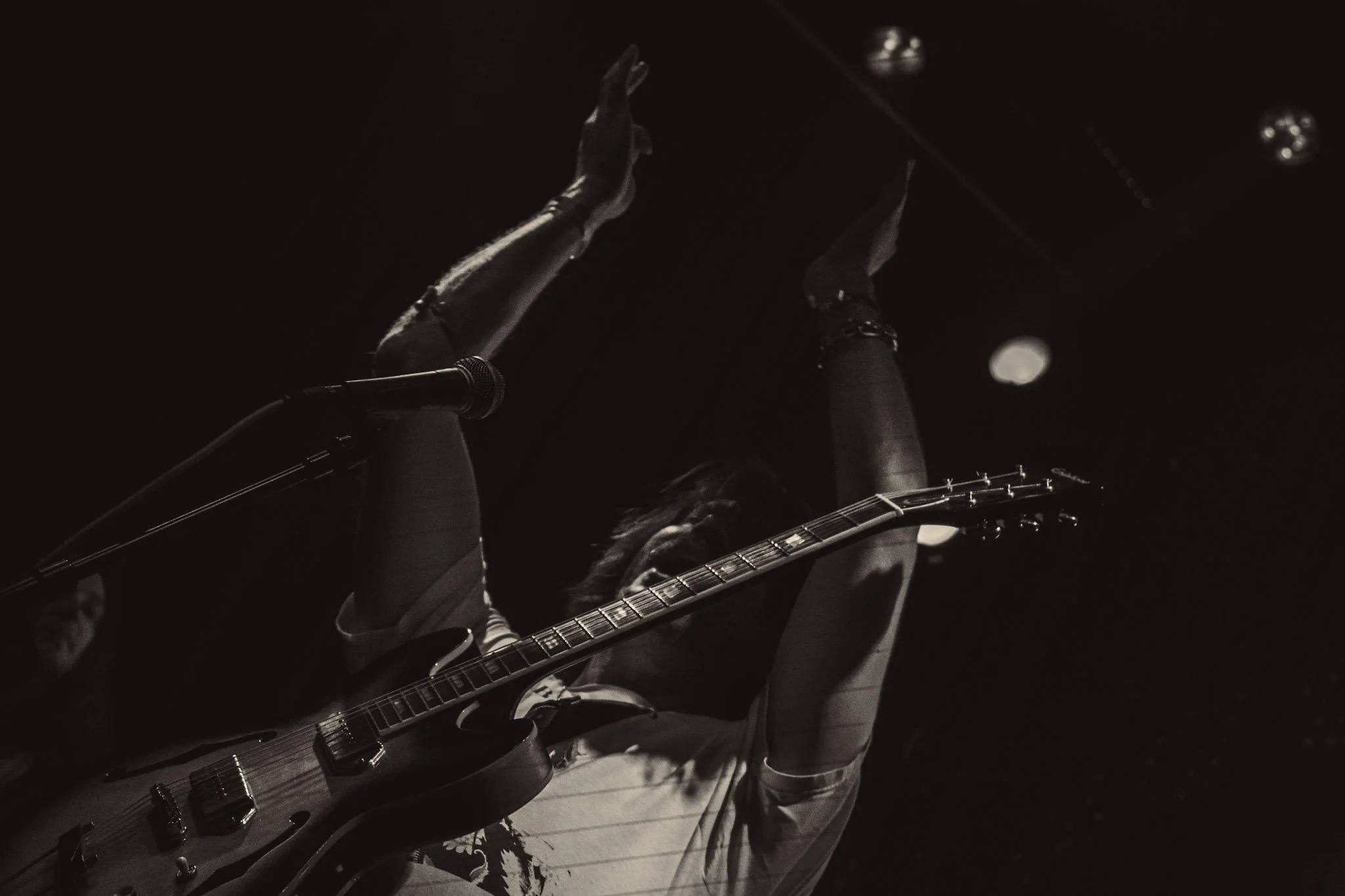 Dark image of a person playing an electric guitar, with their hands raised in the air. The scene is dimly lit, highlighting the guitar and the person's arms.