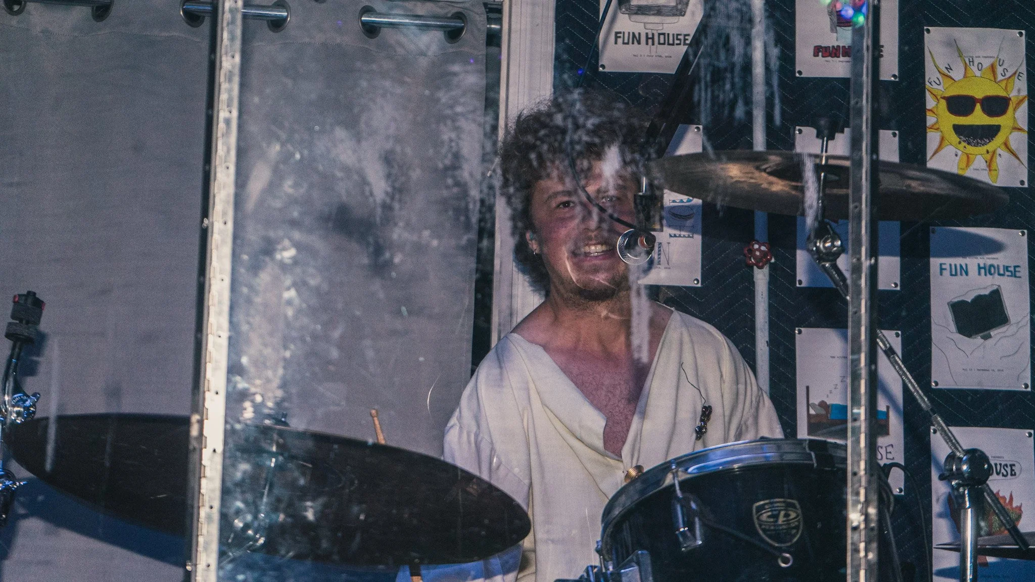 A man with curly hair and a beard, wearing a white shirt, is smiling while sitting behind a drum set in a room with posters labeled 'Fun House'.