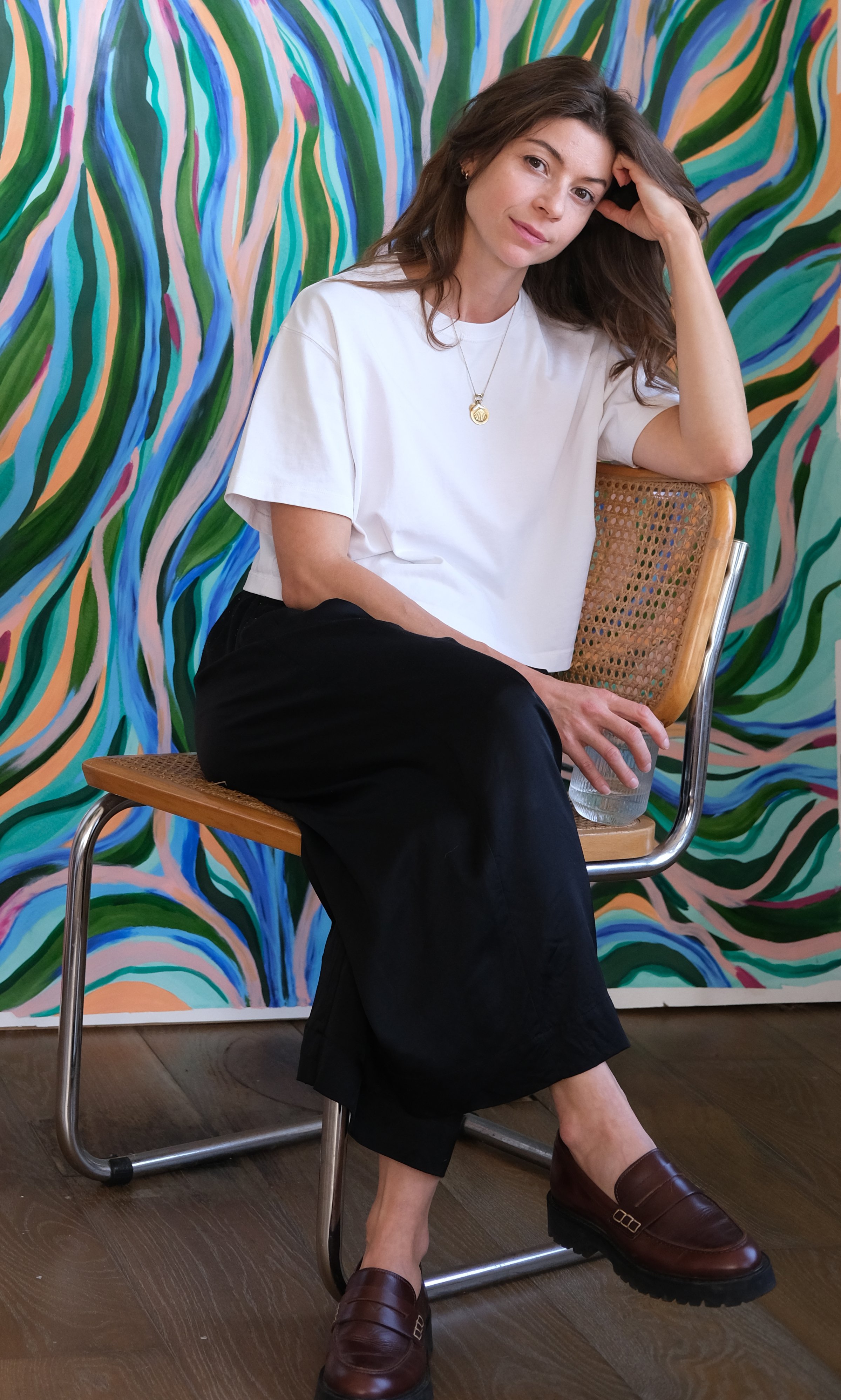 A young woman with long brown hair wearing a white T-shirt, black pants, and brown loafers, sitting on a wooden and cane chair with a colorful, abstract background. She is holding a glass of water and looking at the camera.