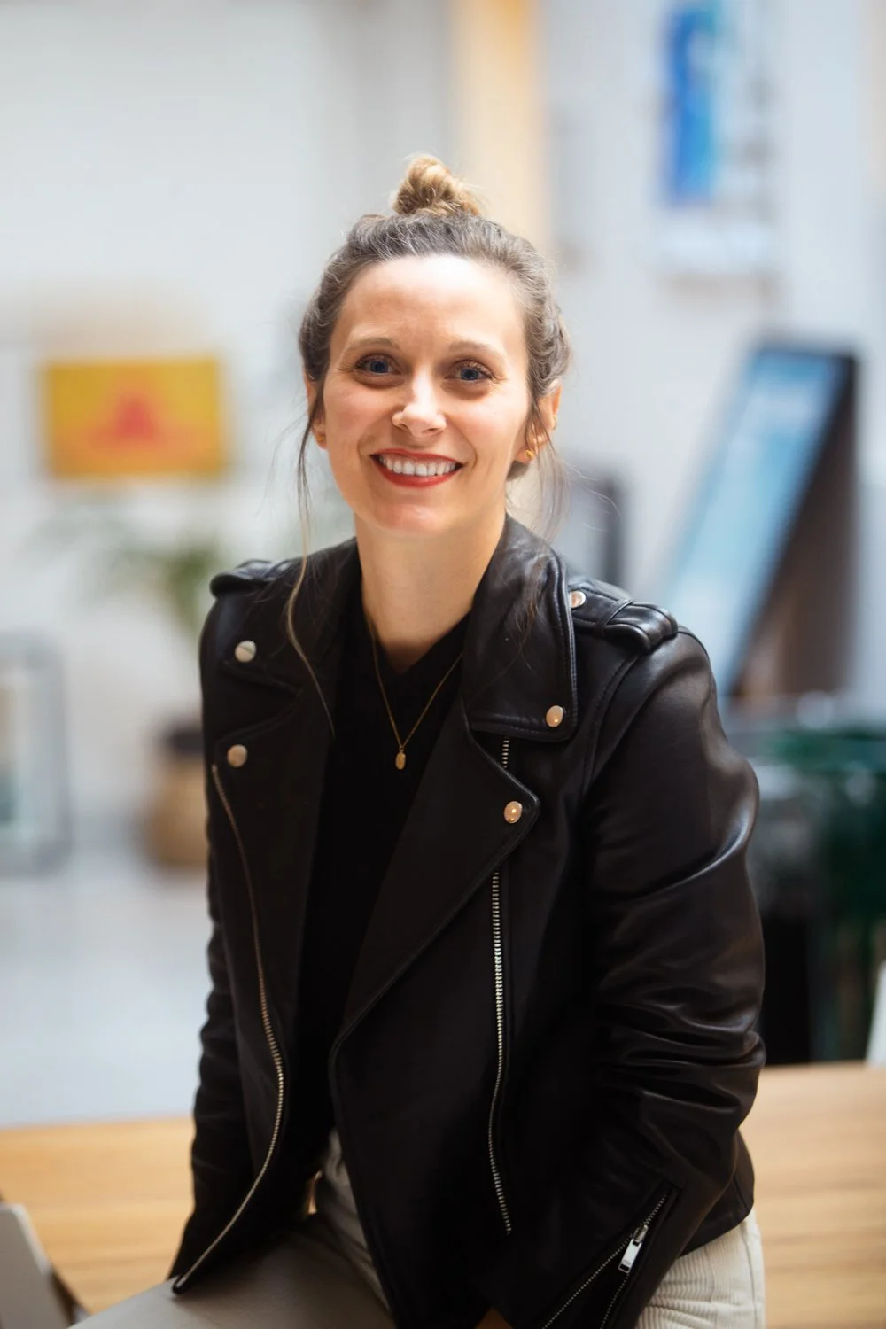A smiling woman with light brown hair in a bun, wearing a black leather jacket and jewelry, sitting at a table in a well-lit office or cafe setting.