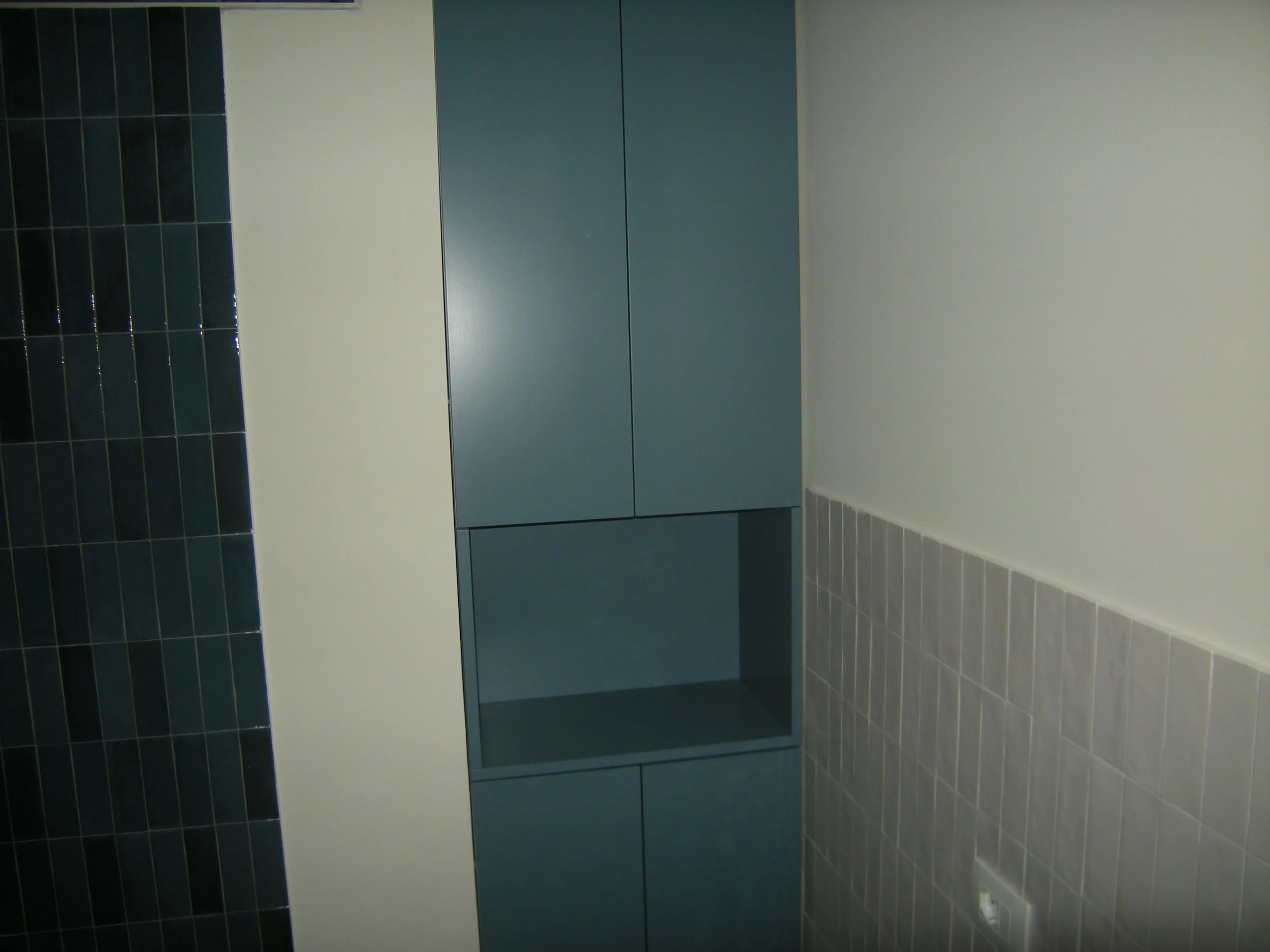 Interno di un bagno con pareti rivestite di piastrelle blu scuro e una parete bianca, mobile bagno blu scuro senza specchio