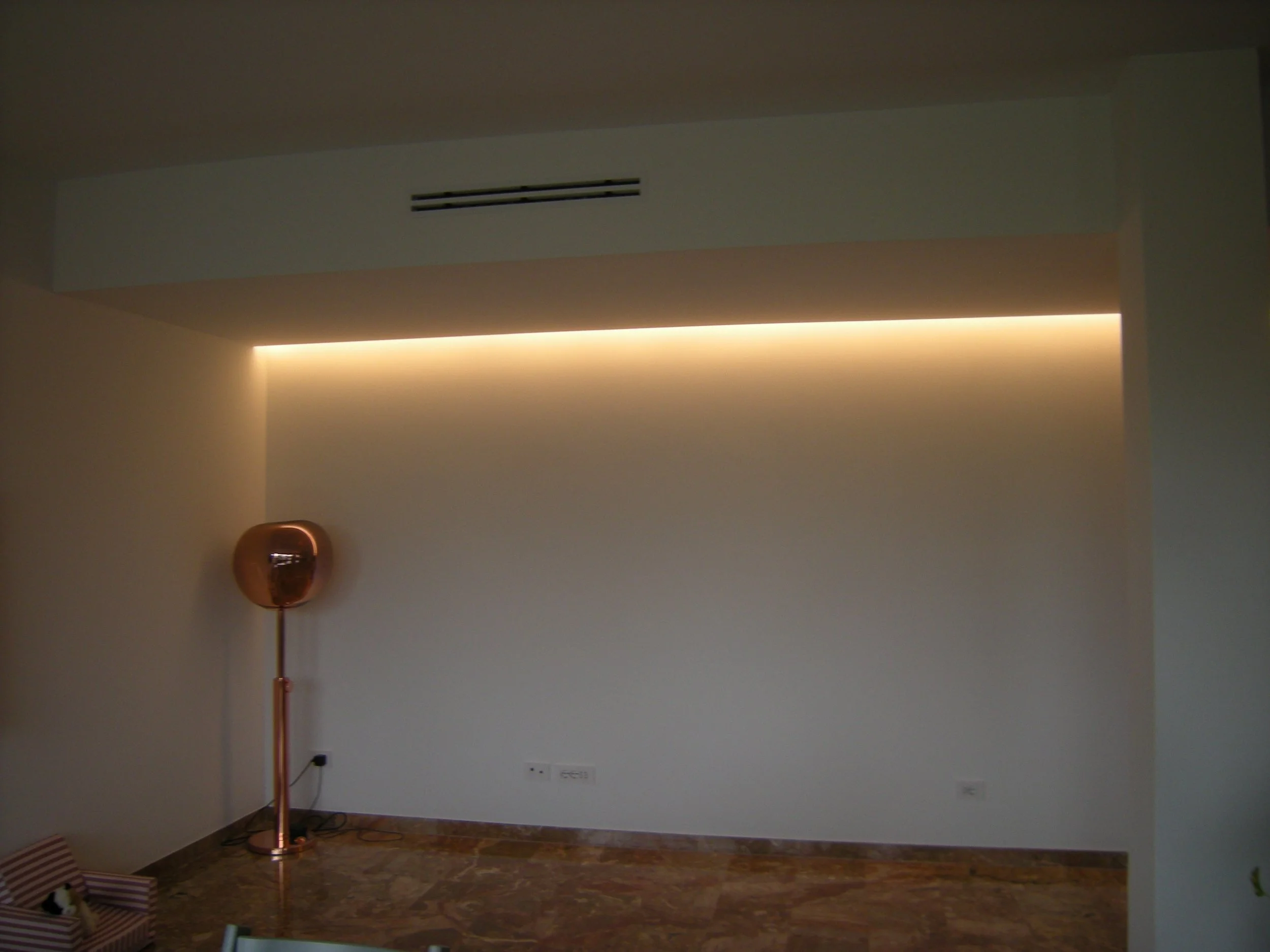 Una stanza vuota con pareti chiare, pavimento di legno e un'illuminazione calda lungo il soffitto. Sul lato sinistro, ci sono una sedia reclinabile a righe e alcuni oggetti sul pavimento.