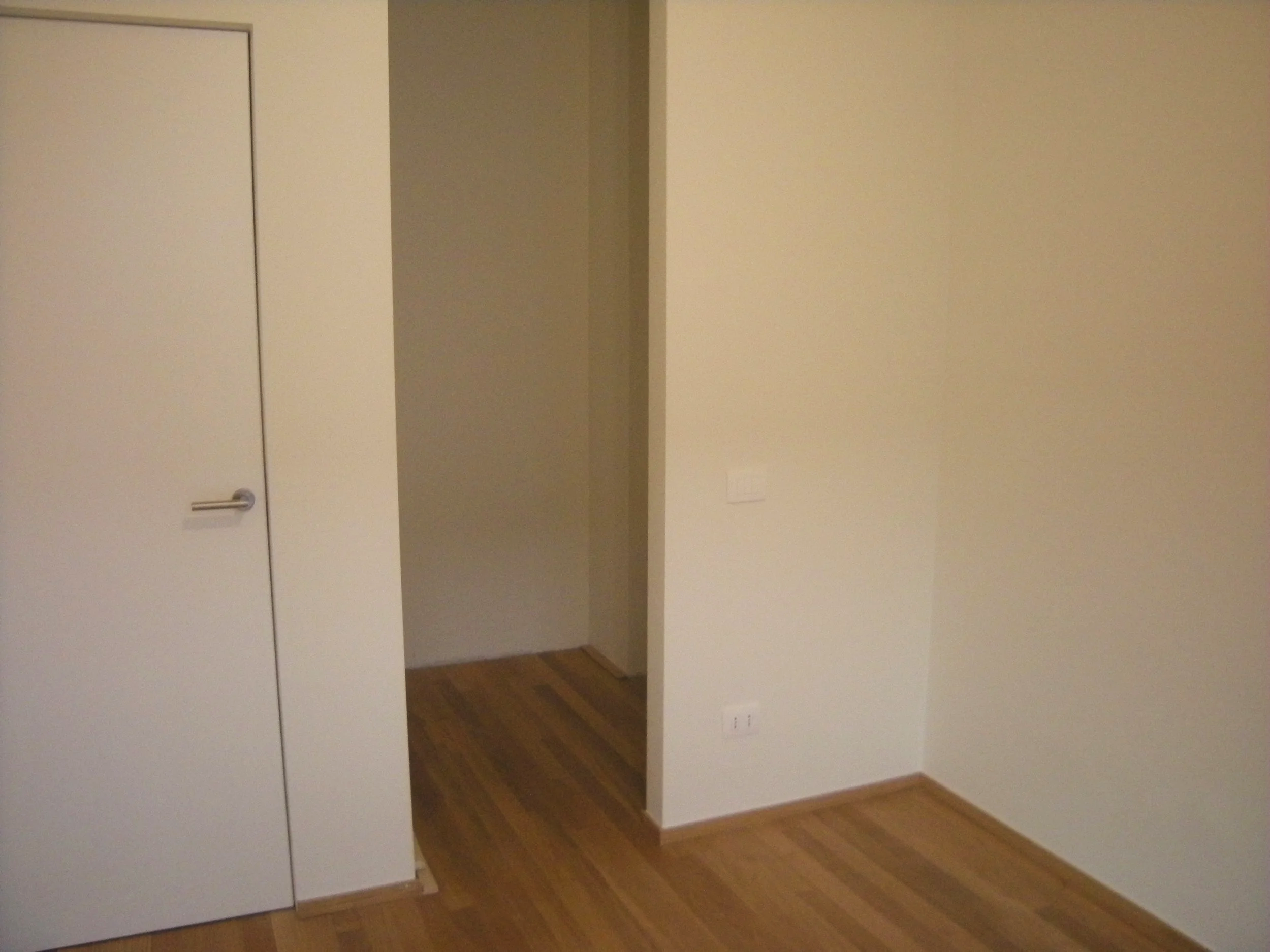 Parete di colore beige con una porta bianca a sinistra e un'apertura senza porta a destra, pavimento in legno e prese elettriche visibili sulla parete.