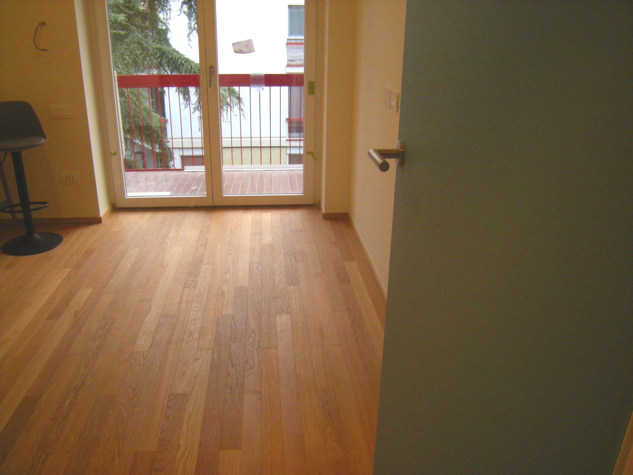Interno di una stanza con pavimento in legno, una porta finestra che apre su un balcone con ringhiera rossa, e una sedia con schienale di fronte alla porta.