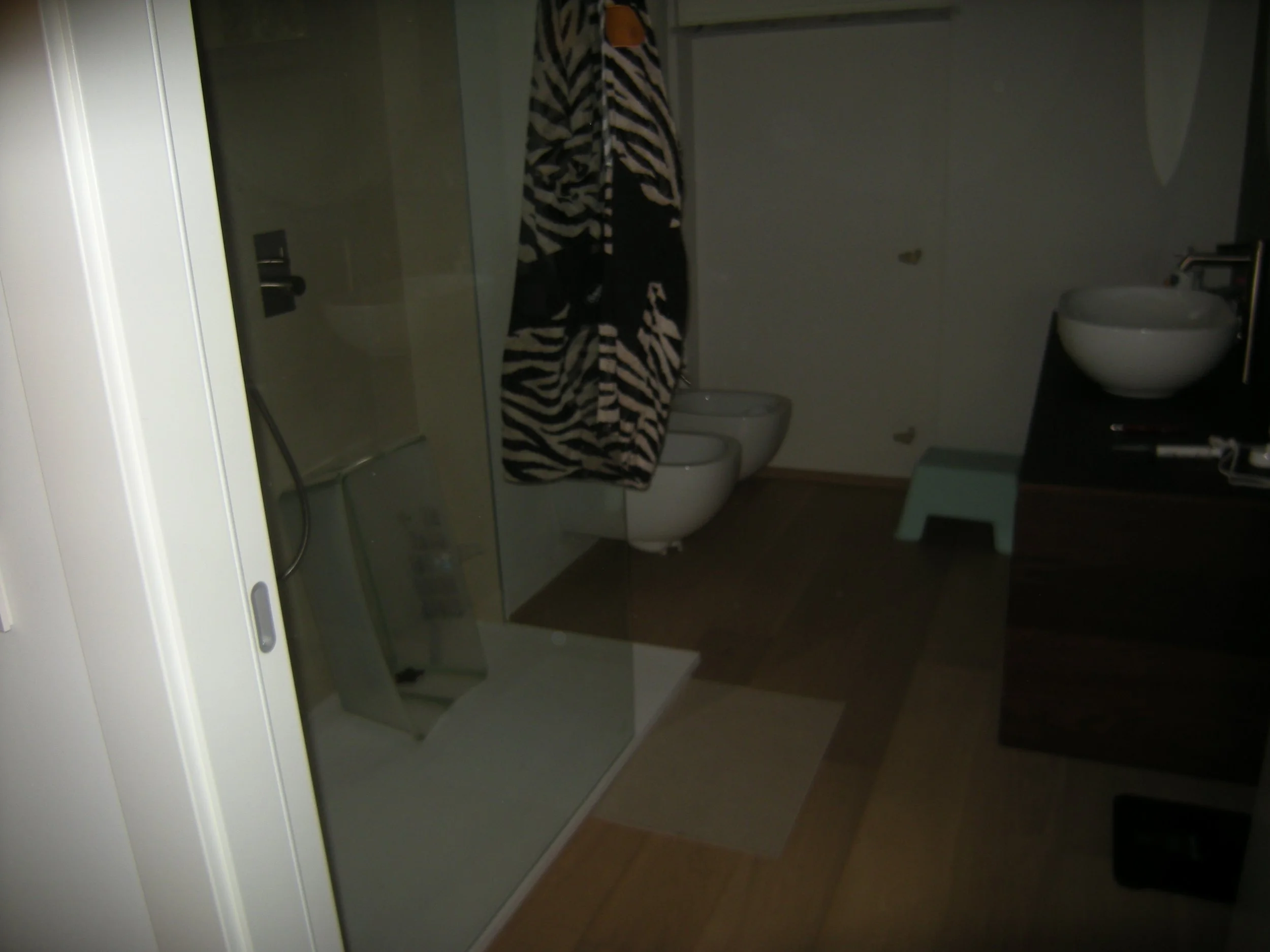 Una stanza da bagno con una cabina doccia, un bidet, un water, una sedia blu e una specchiera. La stanza ha pareti chiare e pavimento in piastrelle di colore beige.