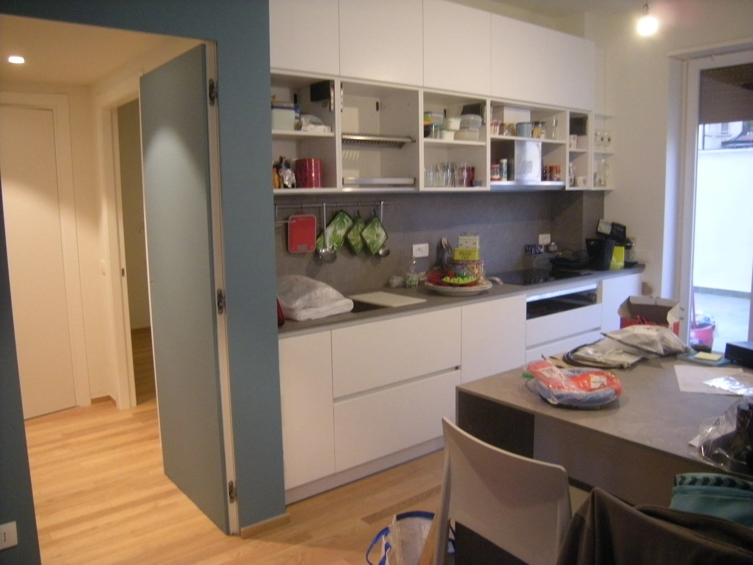 Cucina moderna con scaffali bianchi, mobili bianchi, piano di lavoro con vari oggetti, e una porta finestra che dà su un balcone o terrazza.