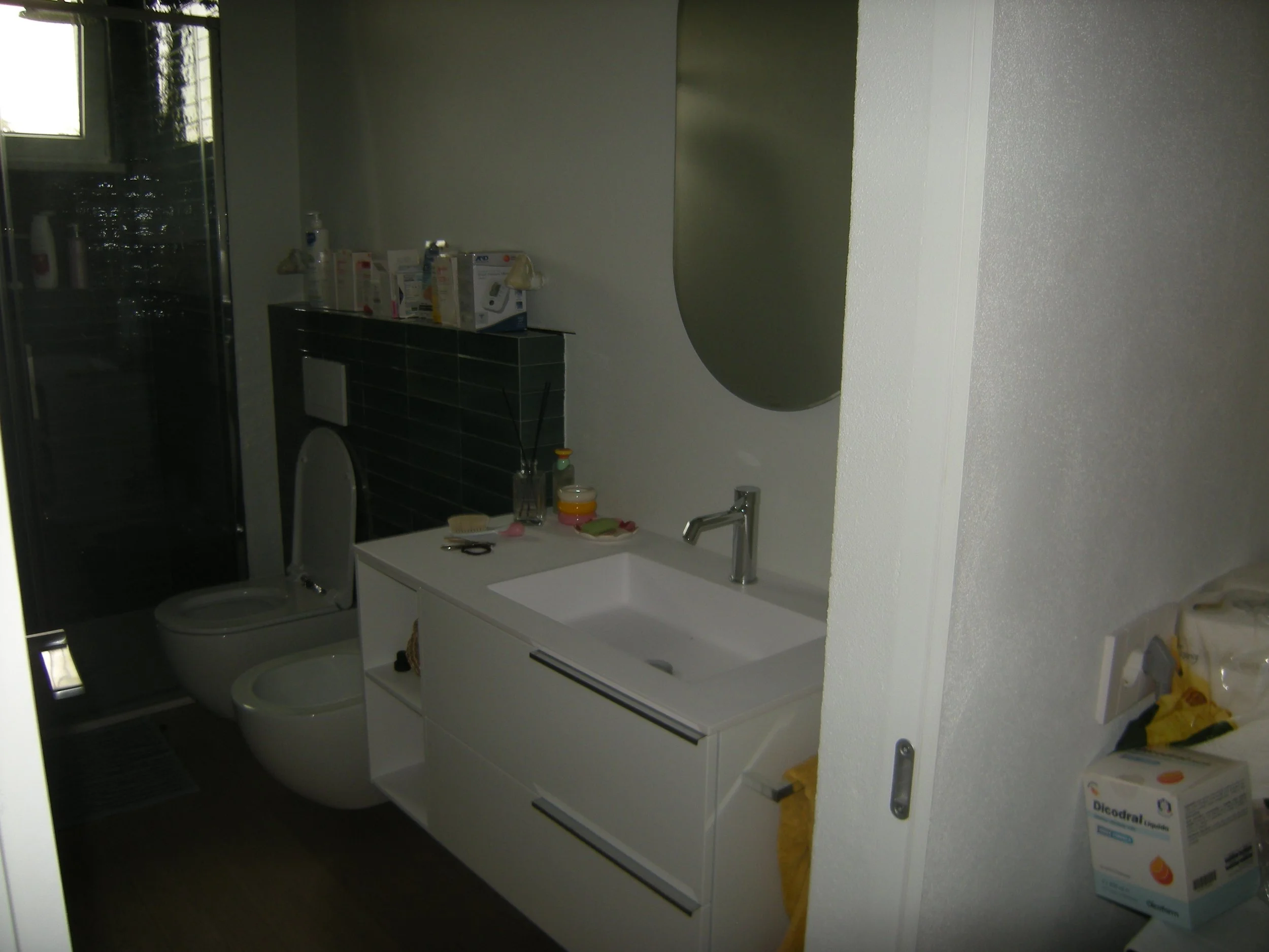 Bagno con lavandino bianco sotto la parete, armadietto bianco con ripiani sopra il lavandino che contiene vari prodotti di cura e alcuni vasetti colorati, toilette in ceramica, cabina doccia con parete in vetro, e scatola di prodotto per la pelle sul