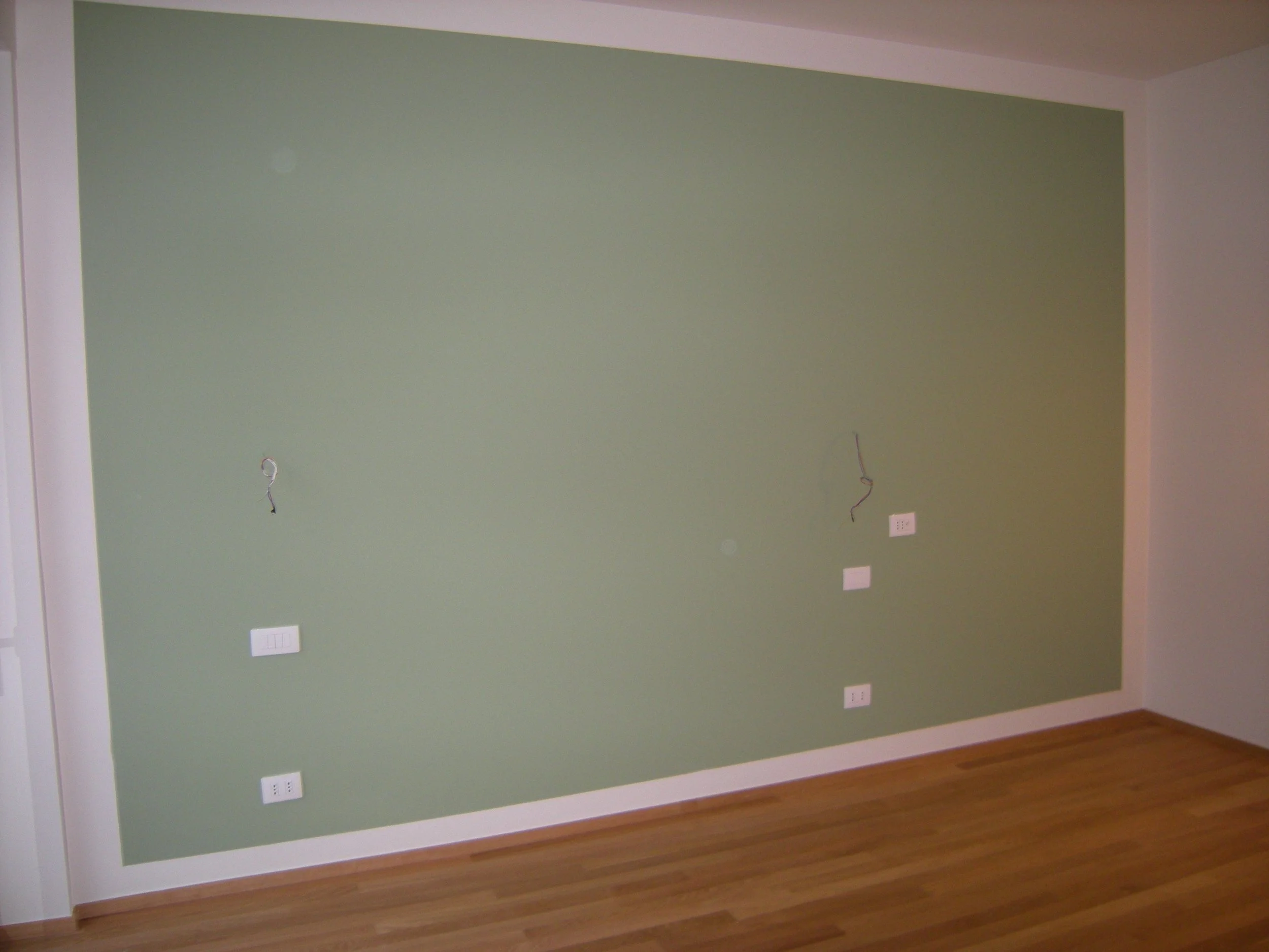 Una parete di colore verde con varia elettronica e prese di corrente lungo il basso della parete in un ambiente domestico o ufficio, con pavimento in legno.