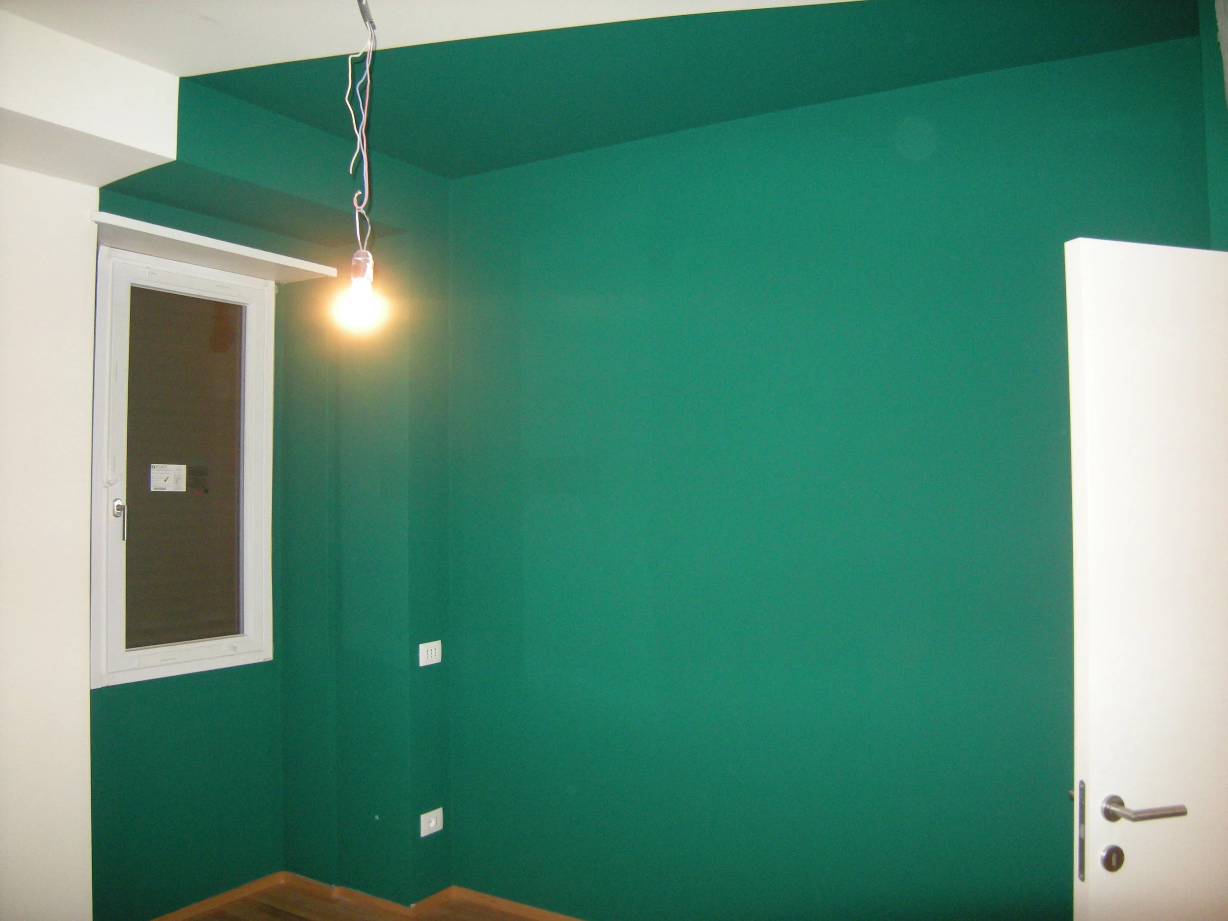 Interno di una stanza con pareti verde scuro, una porta bianca e un pavimento di legno. È presente un errore di installazione con un filo del lampadario pendente dal soffitto, acceso.