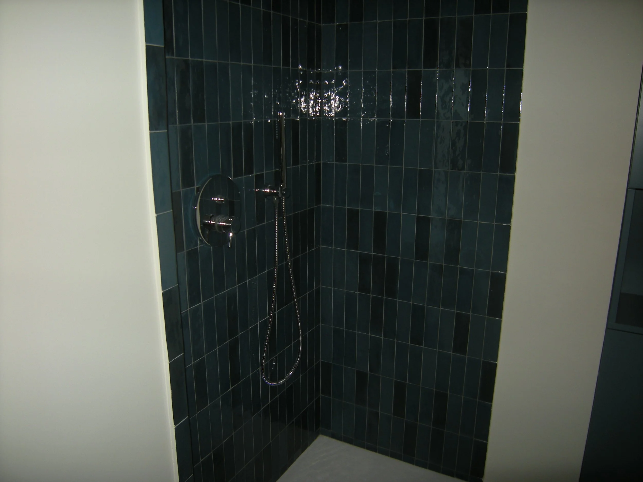 Doccia con piastrelle blu scure, soffitto bianco e un soffione a parete, con flessibile attaccato al soffitto.