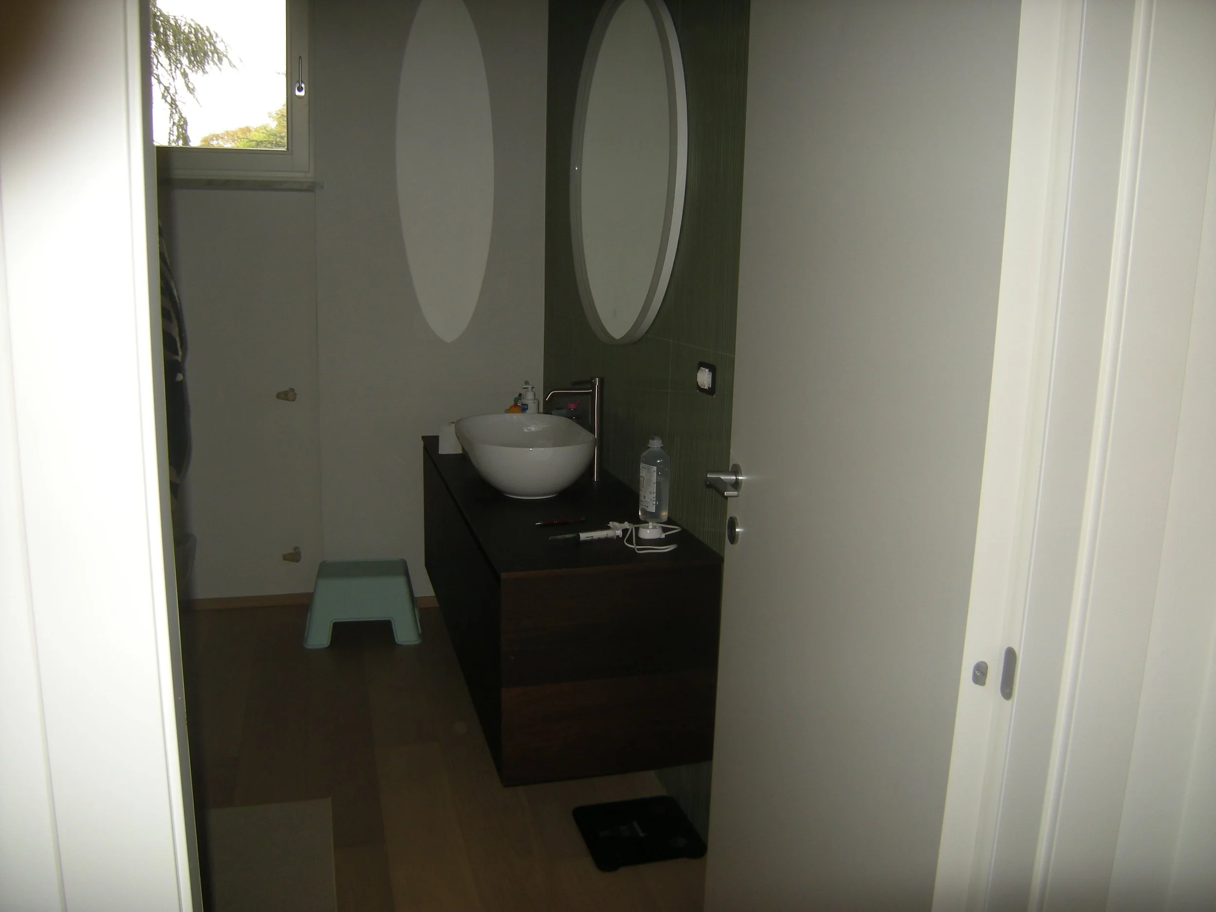 Visuale di un bagno con lavabo rotondo bianco su un mobile scuro, specchio ovale sopra, finestra in alto a sinistra con vista su alberi, panchina bianca sotto il lavabo, pareti chiare, alcuni oggetti sulla superficie del mobile e luci sul soffitto.