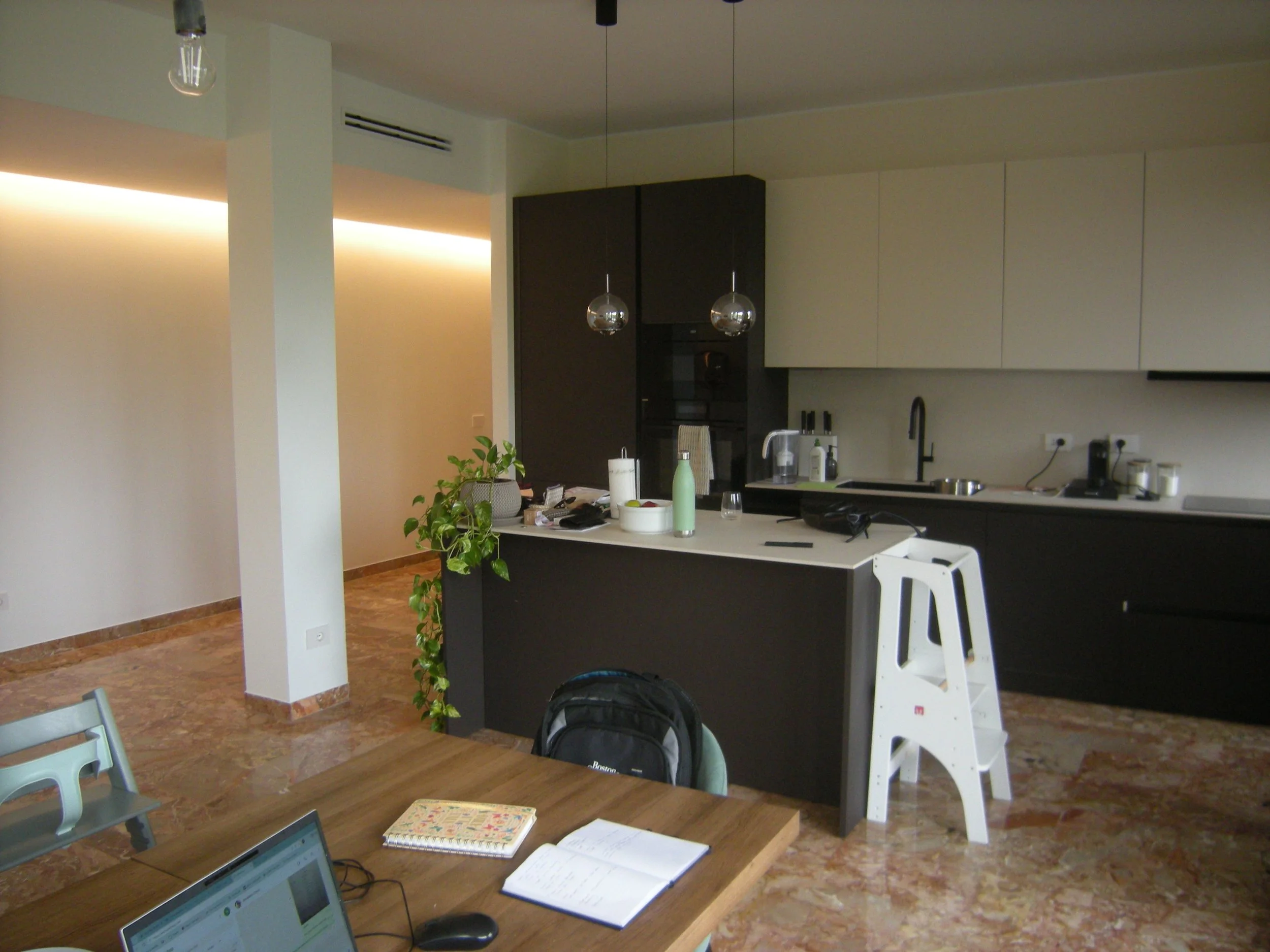 Interno di una cucina moderna con piano di lavoro nero, armadietti bianchi e neri, due lampade a sfera trasparenti, lavello e alcune bottiglie e oggetti sulla superficie, insieme a una pianta verde, una sedia bianca e un tavolo con quaderni e laptop.