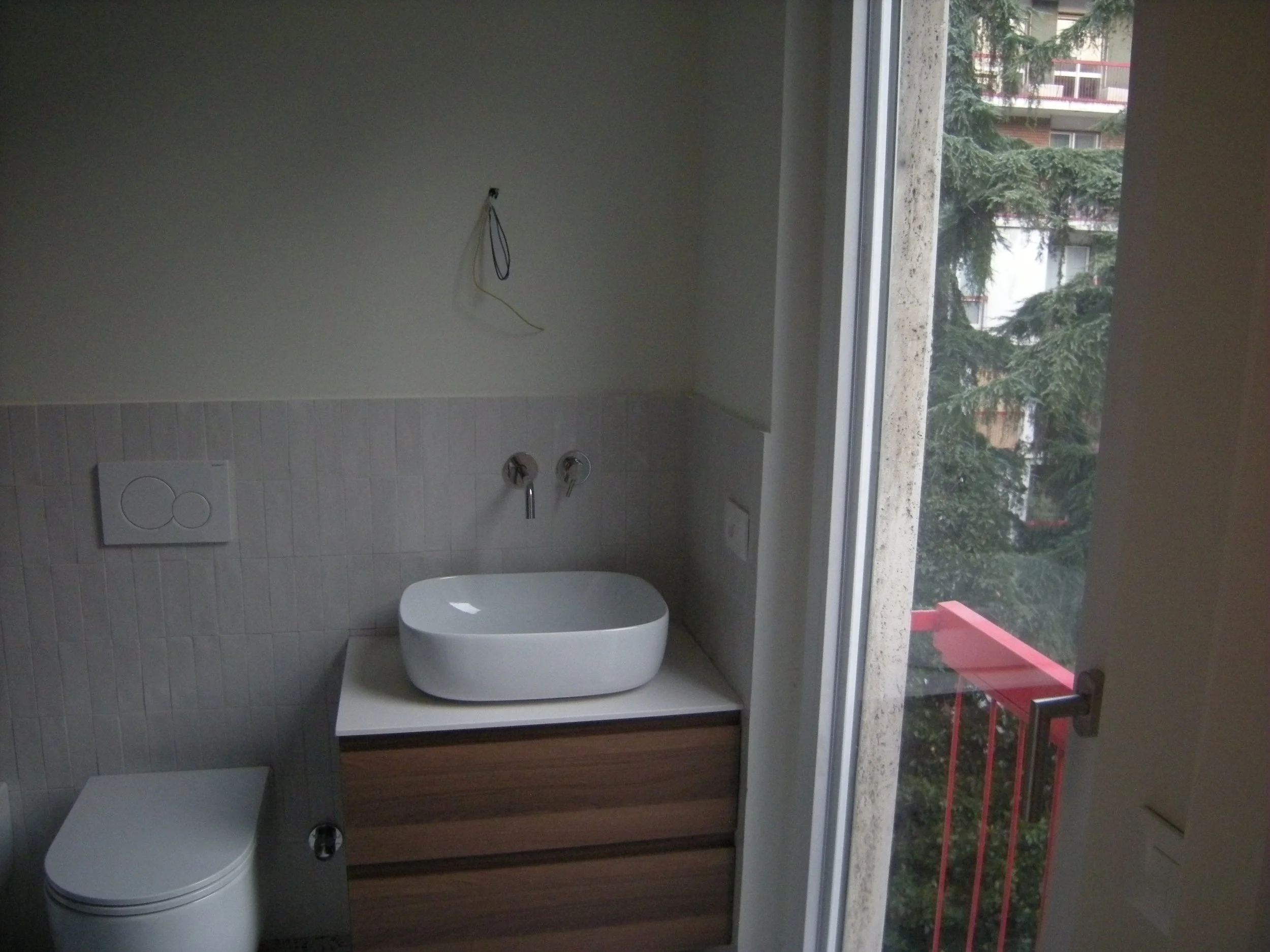 Interno di un bagno con una finestra che si affaccia verso un balcone con tavolo rosso, oltre un albero con rami folti e un edificio con balconi e piante. La stanza ha delle pareti rivestite con piastrelle bianche.