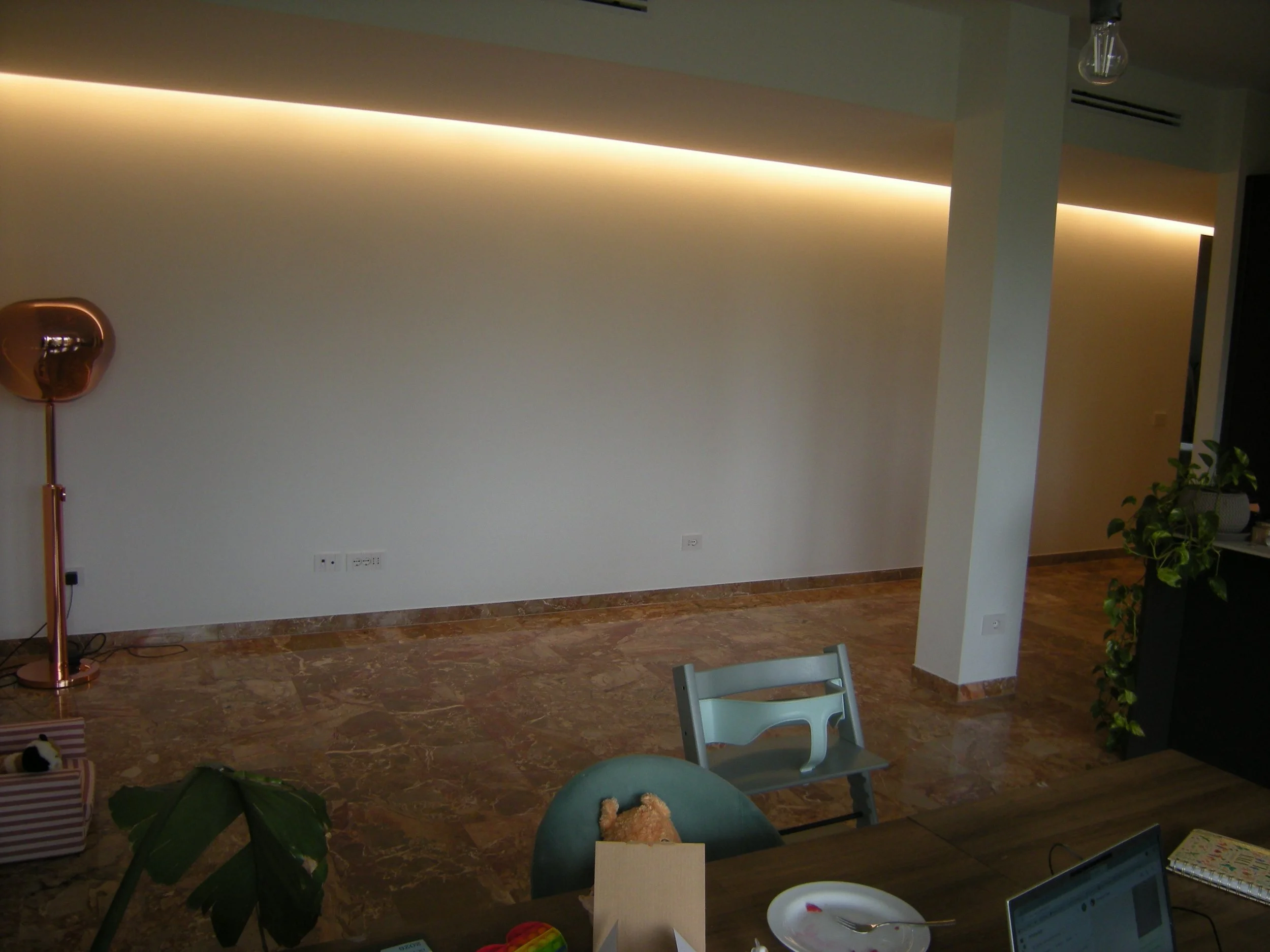 Interno di una stanza con pareti bianche e soffitto con illuminazione incorporata, pavimento in marmo, presenza di piante e mobili, incluso un tavolo con sedie colorate.