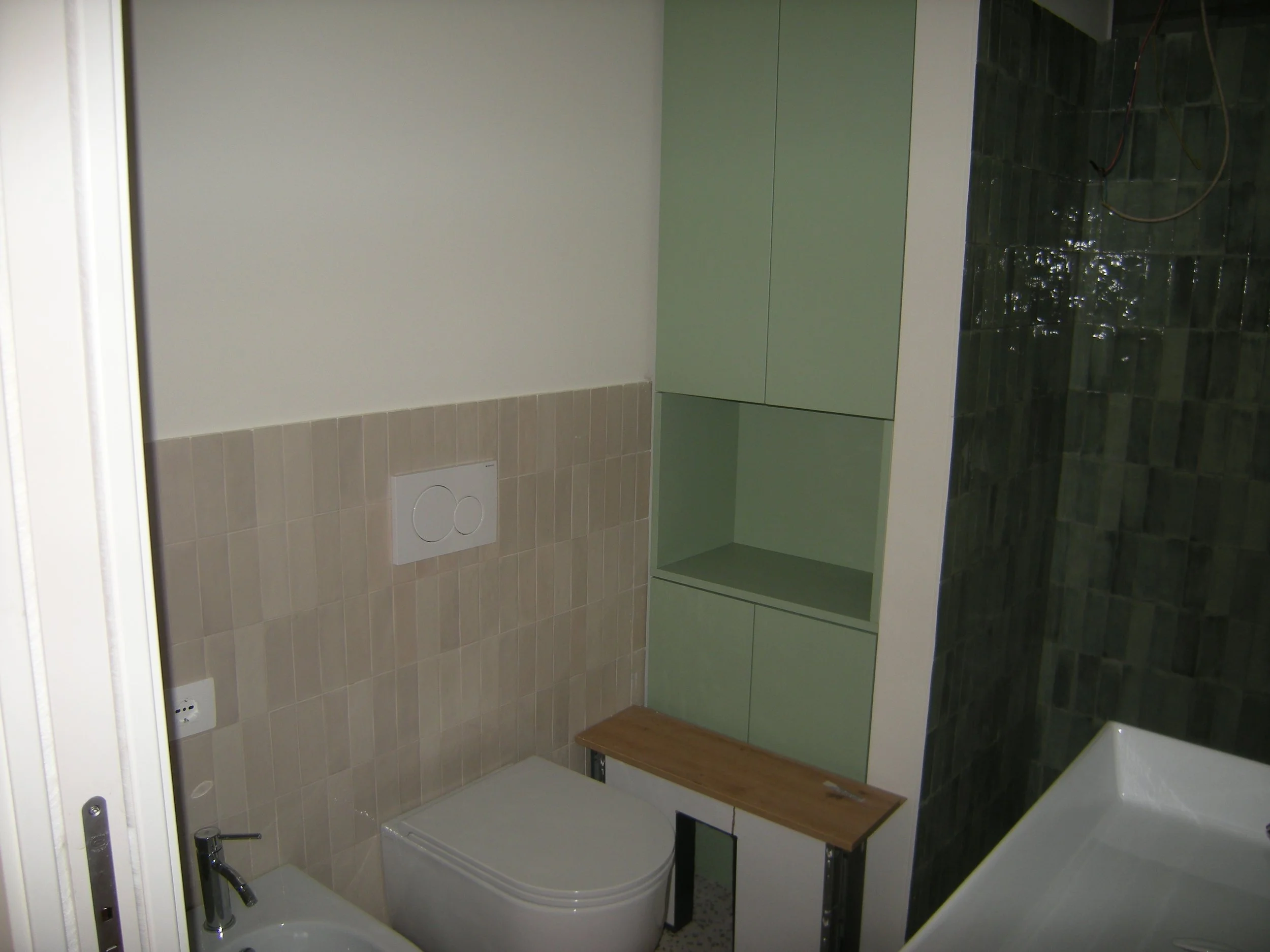 Interno di un bagno con pareti in piastrelle beige, armadietto verde e sanitari con vaso e bidet, pavimento in pietra. C'è una finestra e un interruttore di corrente.