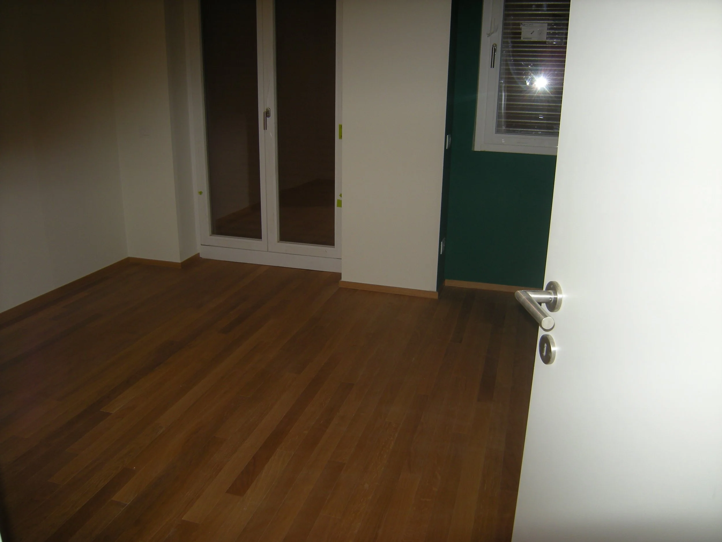 Stanza vuota con pareti bianche e verdi, pavimento in legno, finestra con tapparella abbassata, porta aperta con maniglia in metallo.