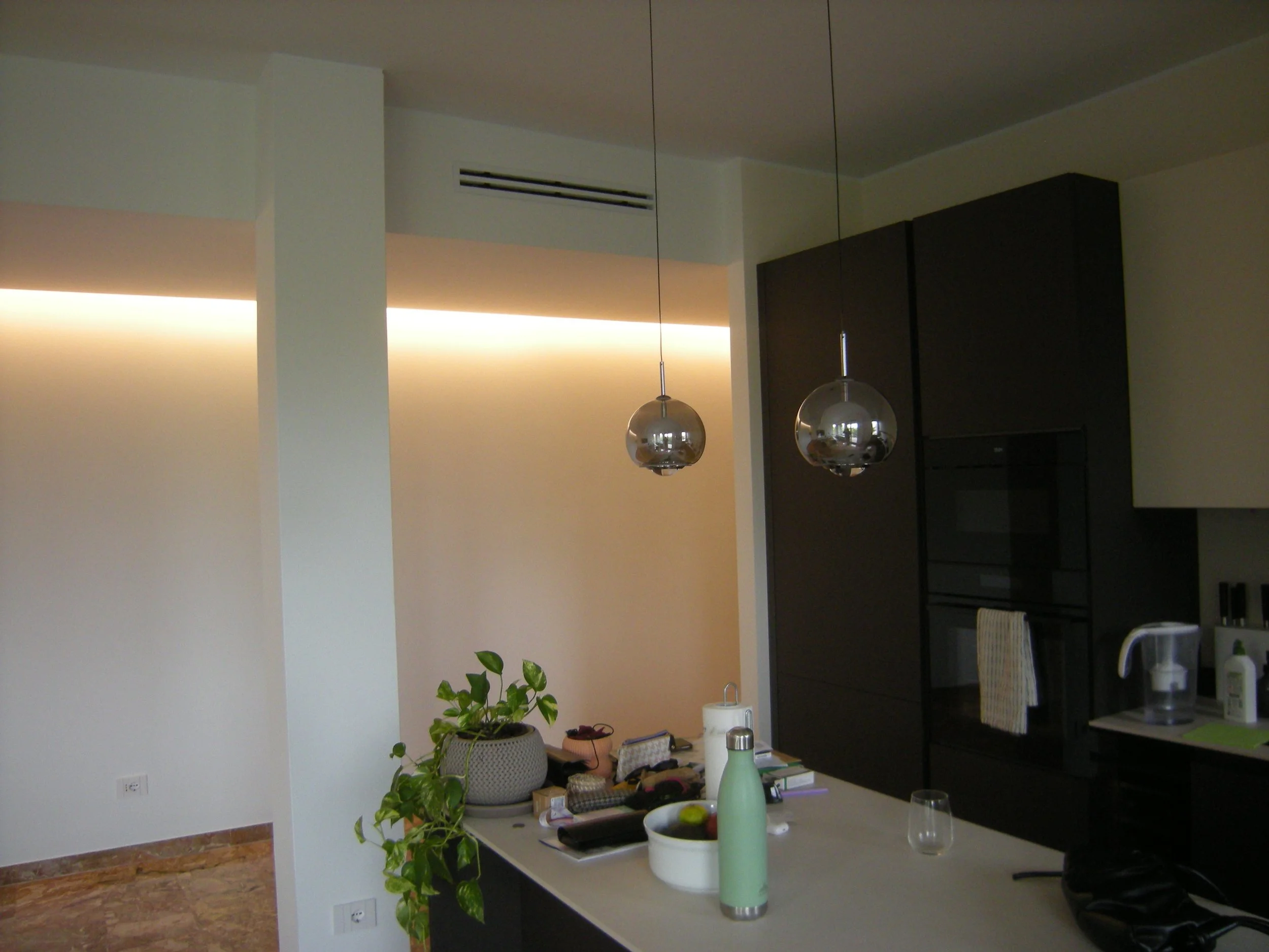 Interno di una casa con zona pranzo e cucina, piante, luci a soffitto e vari oggetti sulla superficie.