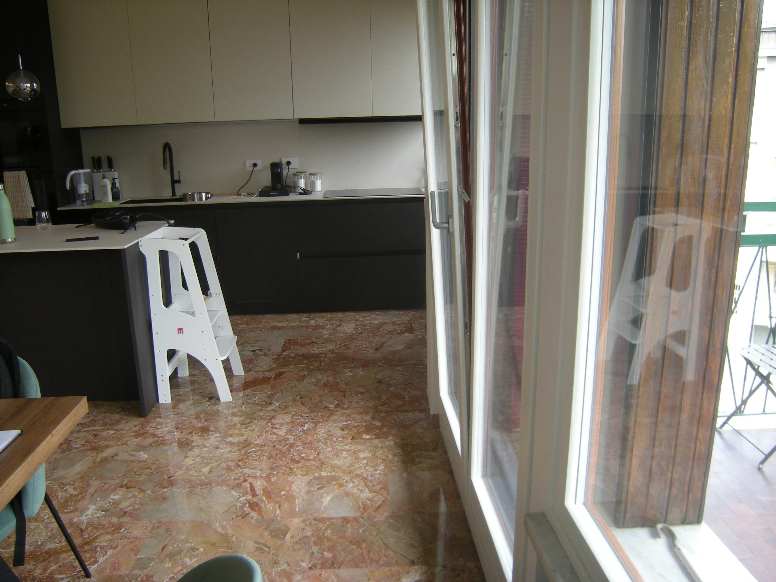 Interno di una cucina con un piano di lavoro, armadietti beige sopra un piano nero, una finestra a vetri dalla quale si vede un balcone con pavimento in legno, e una parete con mattoni rossi.