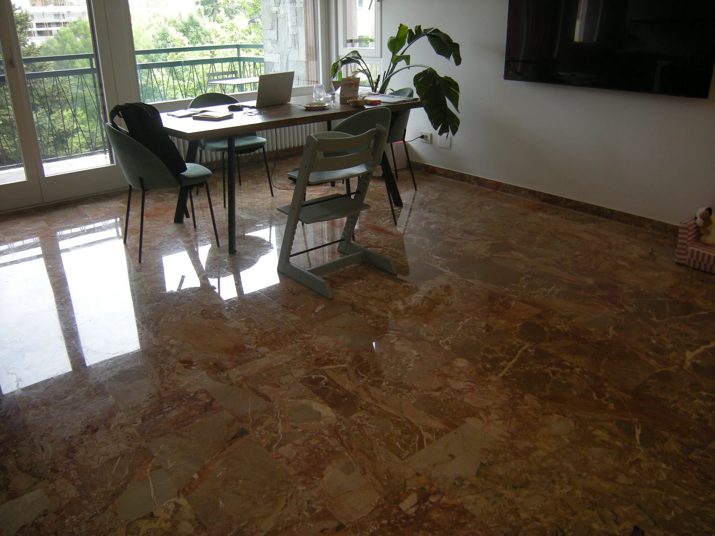 Interno di una stanza con un tavolo di legno, sgabelli, una sedia alta e una pianta grande vicino alla finestra.
