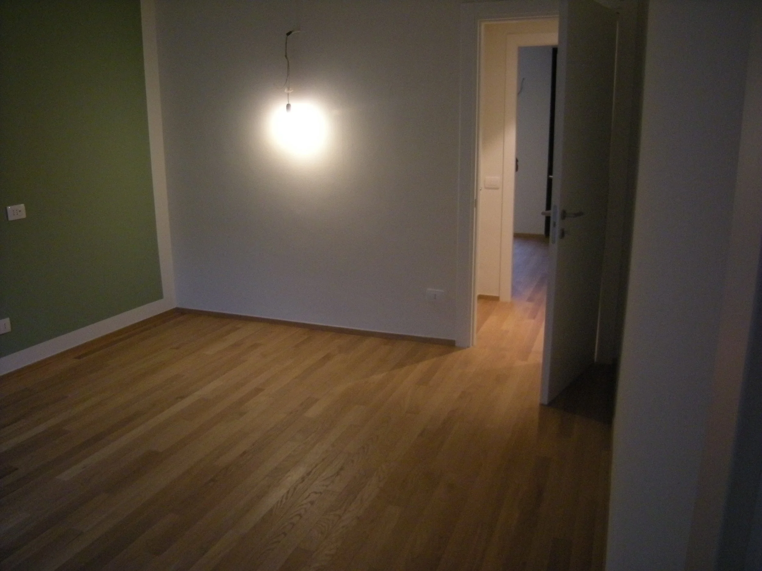 Stanza vuota con pareti di colore grigio e verde, pavimento di legno, una porta aperta e luce da plafoniera