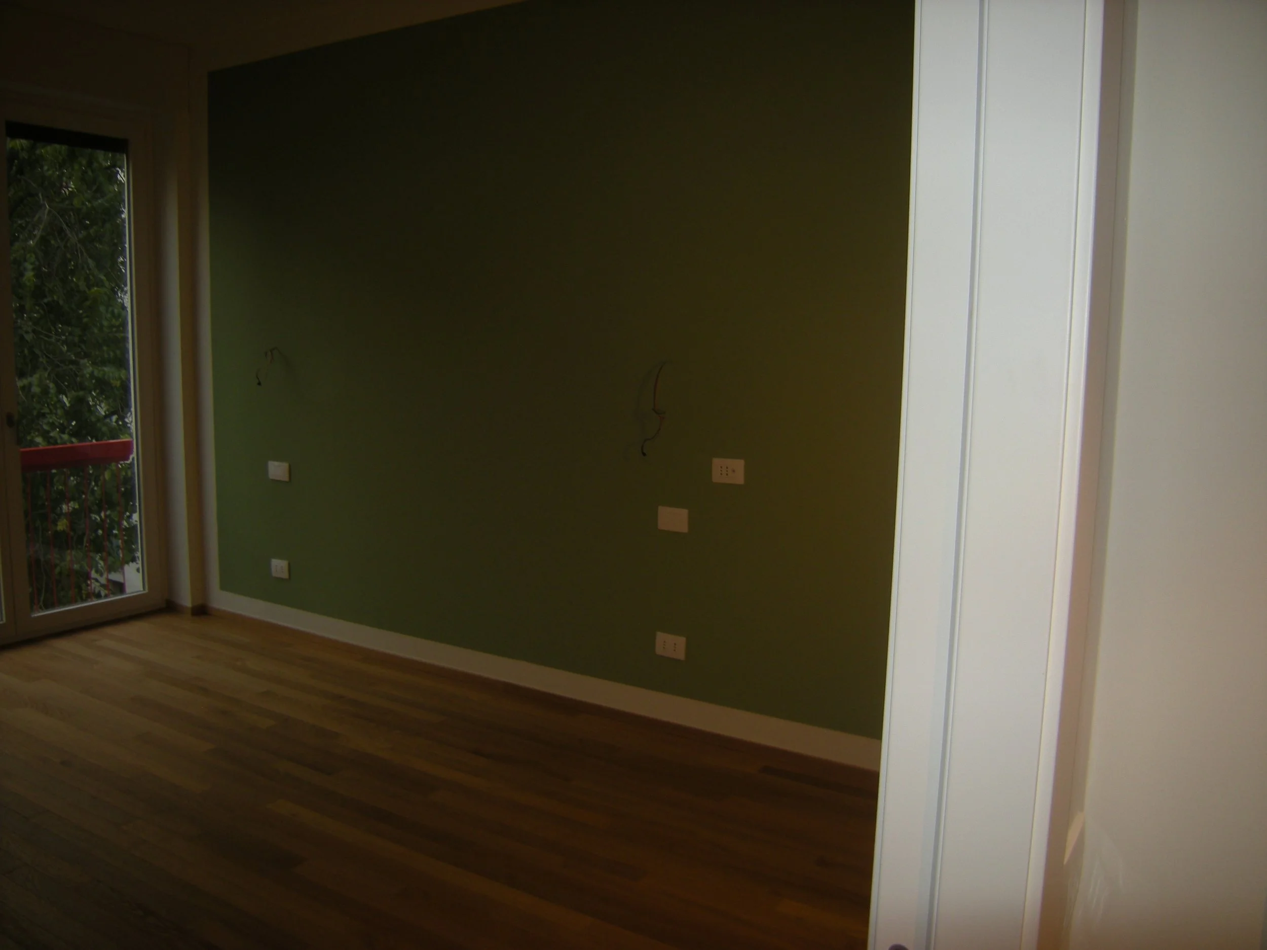 Interno di una stanza con pareti verde scuro e bianca, prese di corrente e cablaggio visibile, pavimento in legno