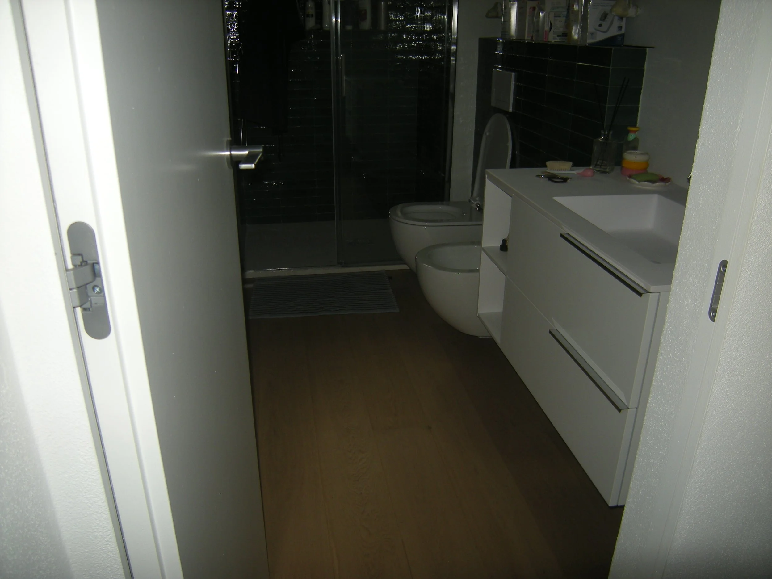 Interno di un bagno con lavabo bianco, mobile a due cassetti, specchio e vari oggetti come una bottiglia di sapone, un portasciugamano, e altri accessori.