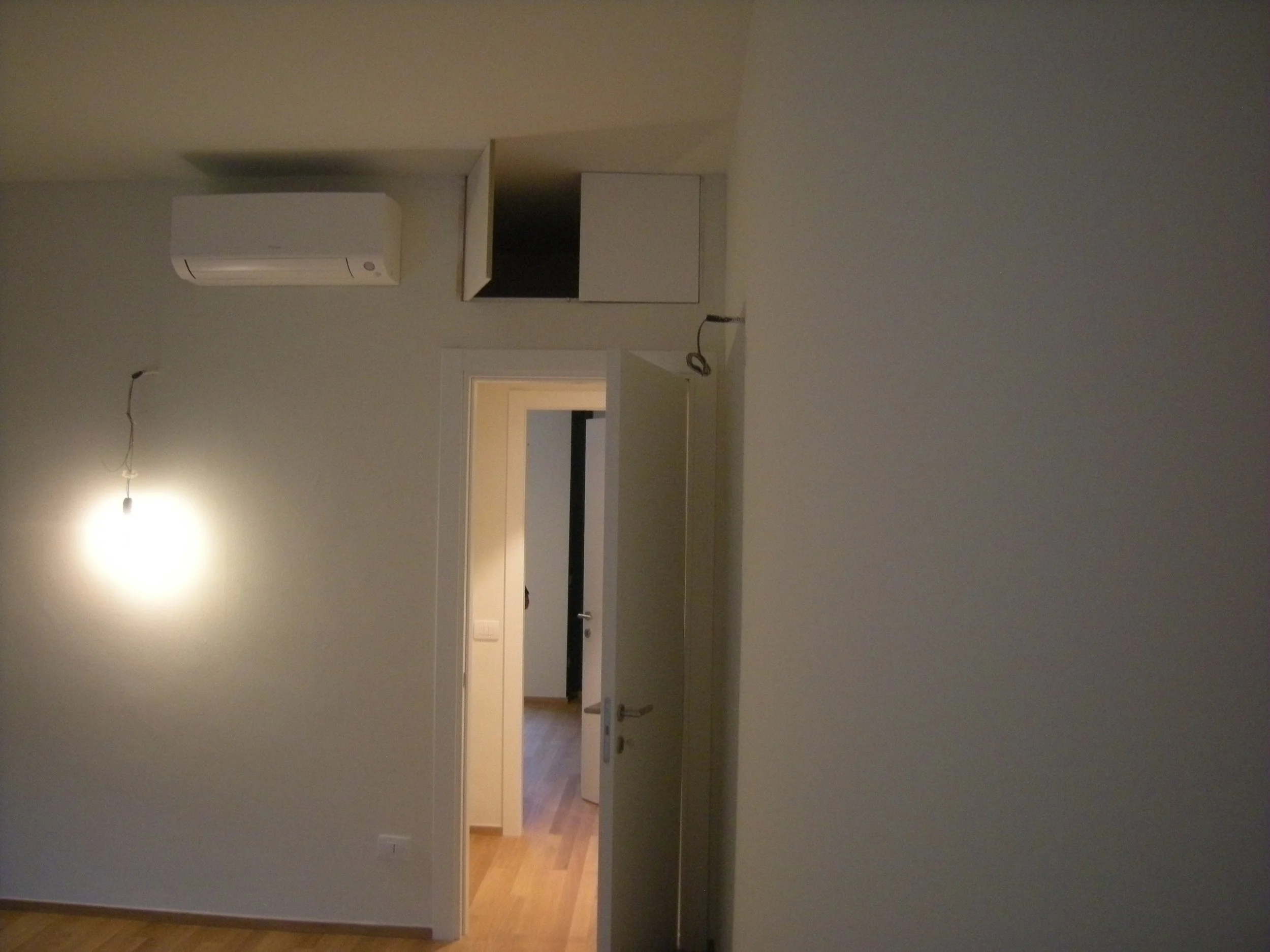 Immagine di una stanza con pareti verde chiaro, una porta aperta che mostra un'altra stanza con pavimento in legno e pareti beige. Sul soffitto si vede una luce spenta e un condizionatore bianco sulla parete.