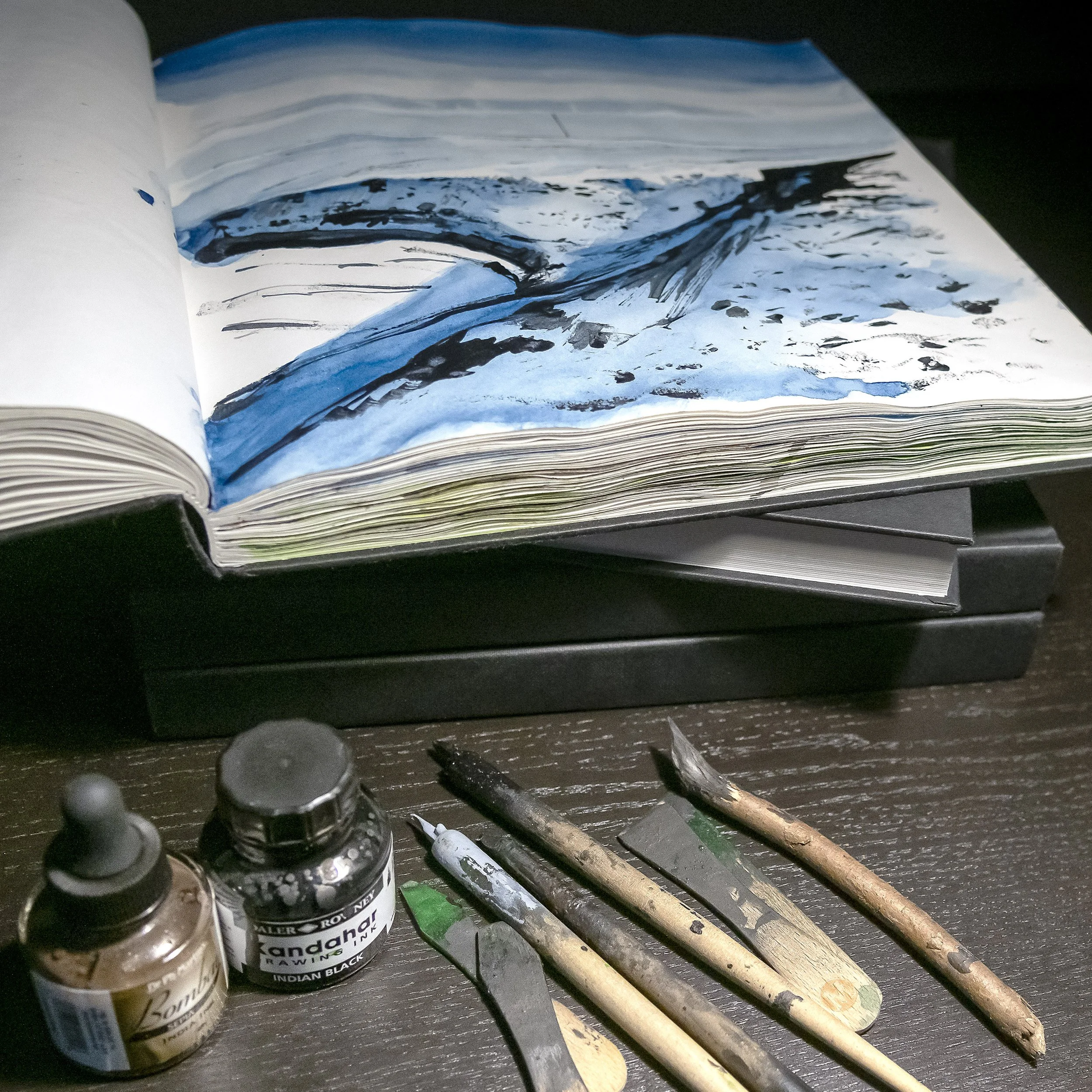 Open sketchbook with a blue and black abstract painting, surrounded by paintbrushes and ink bottles on a worktable.