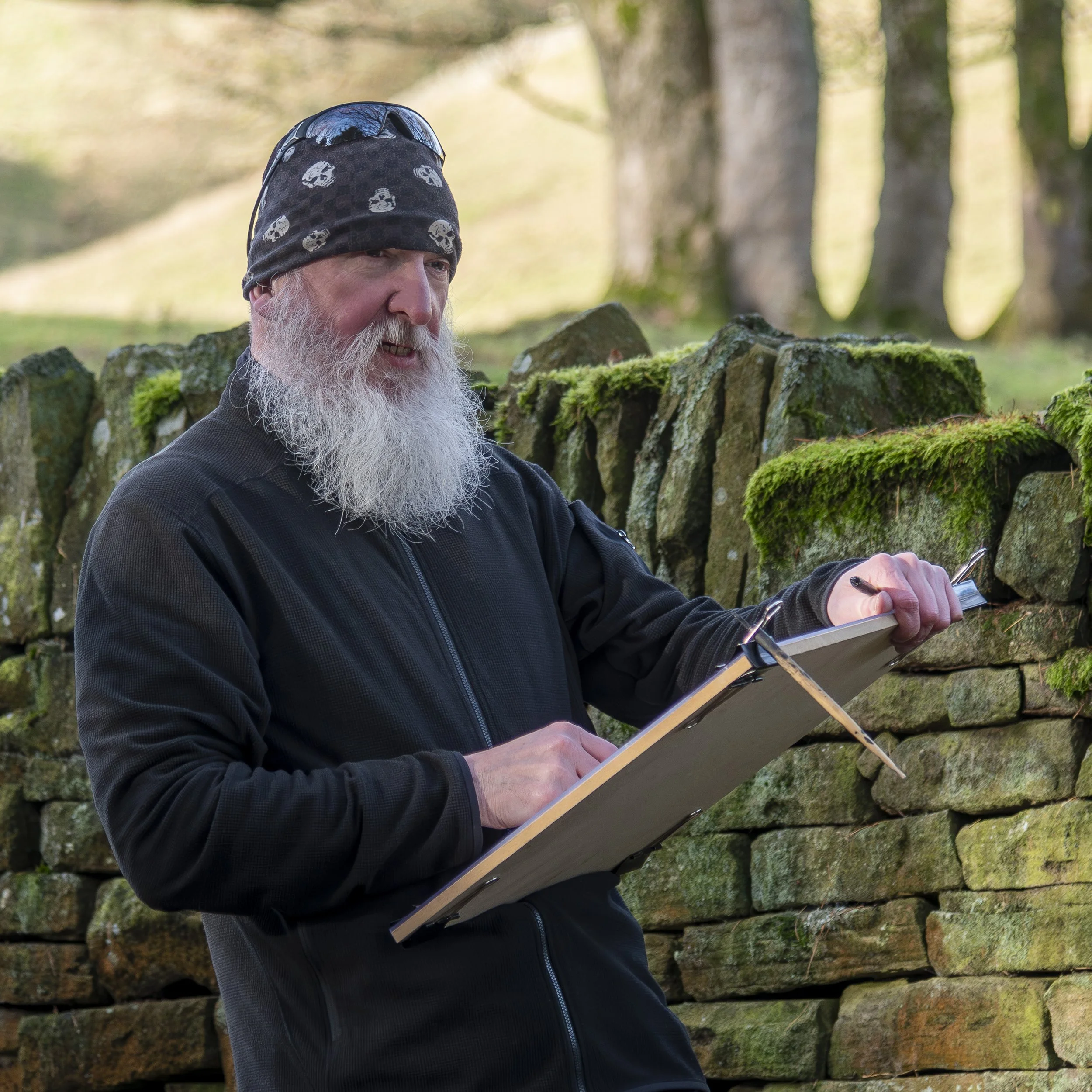 Bearded man wearing a skull-patterned beanie and sunglasses, writing on a clipboard beside a moss-covered stone wall in the woods