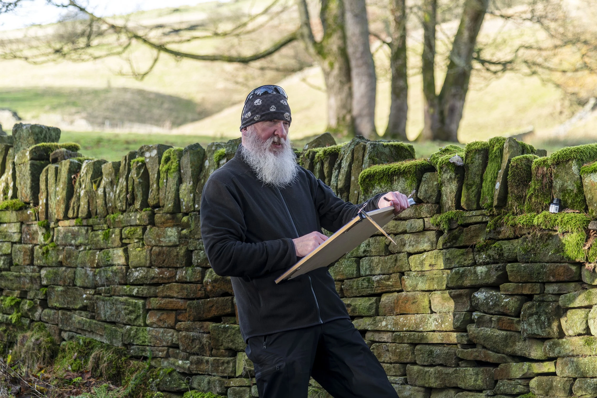 Bearded man wearing a skull-patterned beanie paints outdoors beside a moss-covered stone wall.