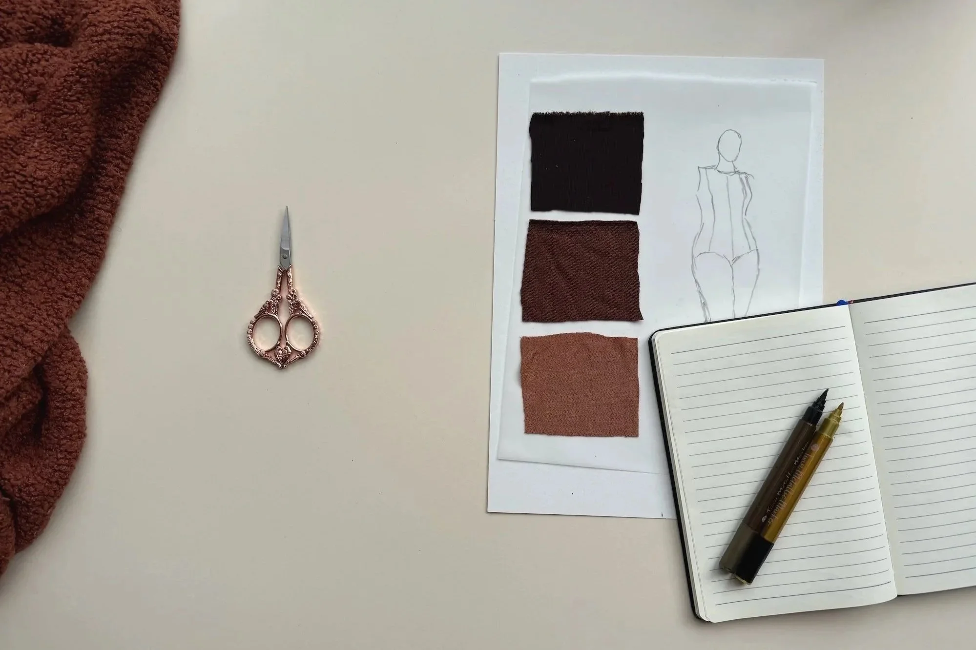 Sewing scissors beside fabric swatches and a notebook with clothing sketches.