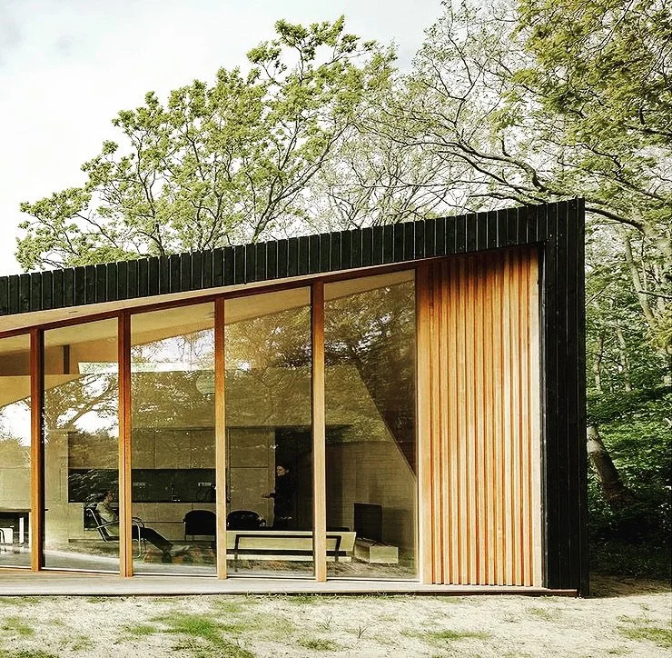 Modern house with large glass windows and black and wooden exterior, surrounded by trees.