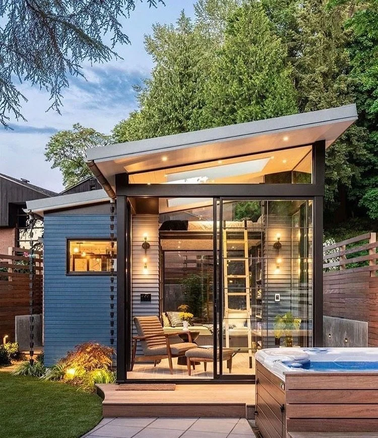 Modern glass-walled backyard tiny house with outdoor seating and hot tub, surrounded by greenery and wooden fencing.