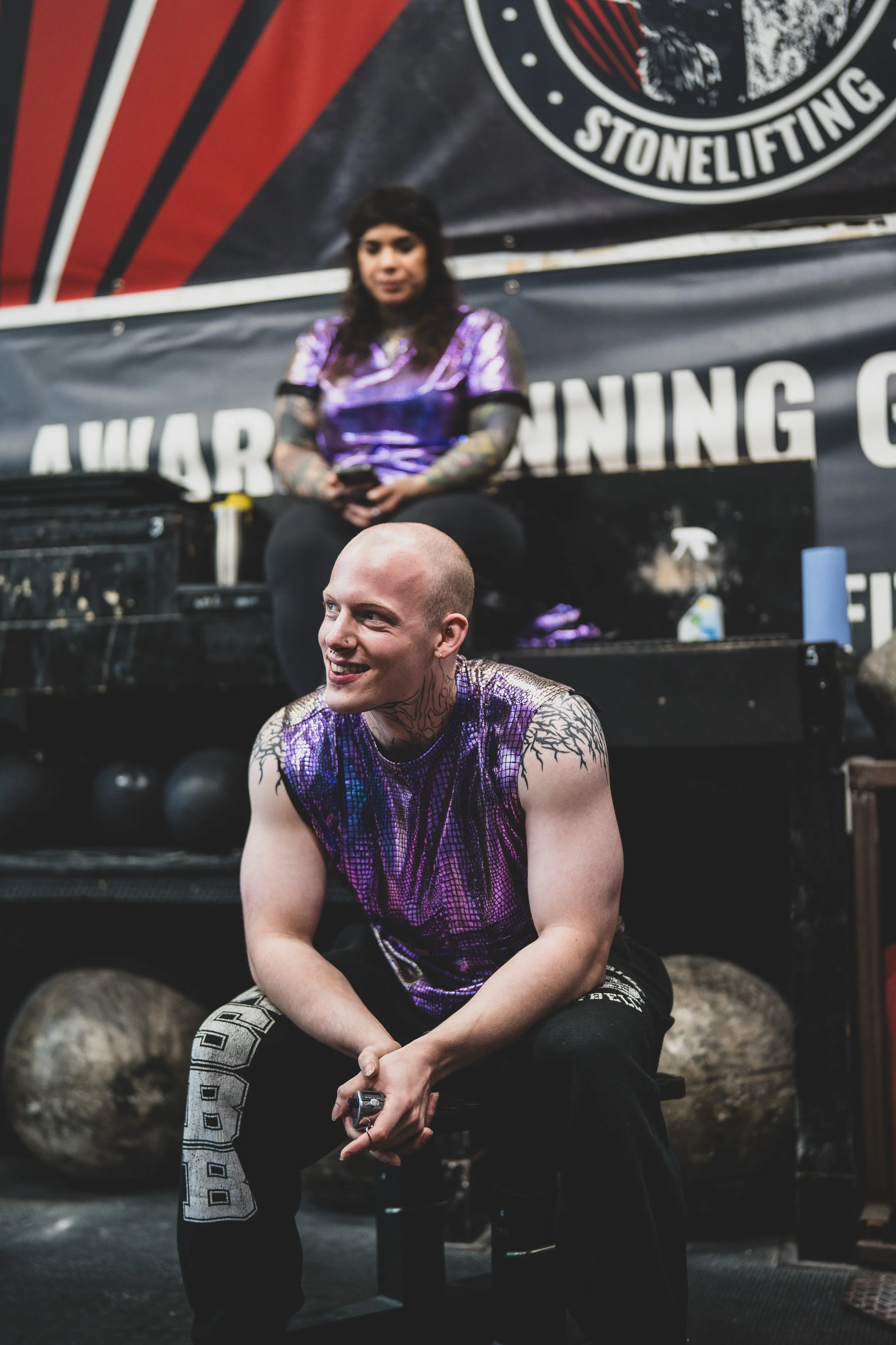 Two muscular individuals with tattoos, wearing purple and black workout clothing, seated in a gym with stone lifting weights and a banner that reads 'AWARD WINNING G...' and 'STONELIFTING' in the background.