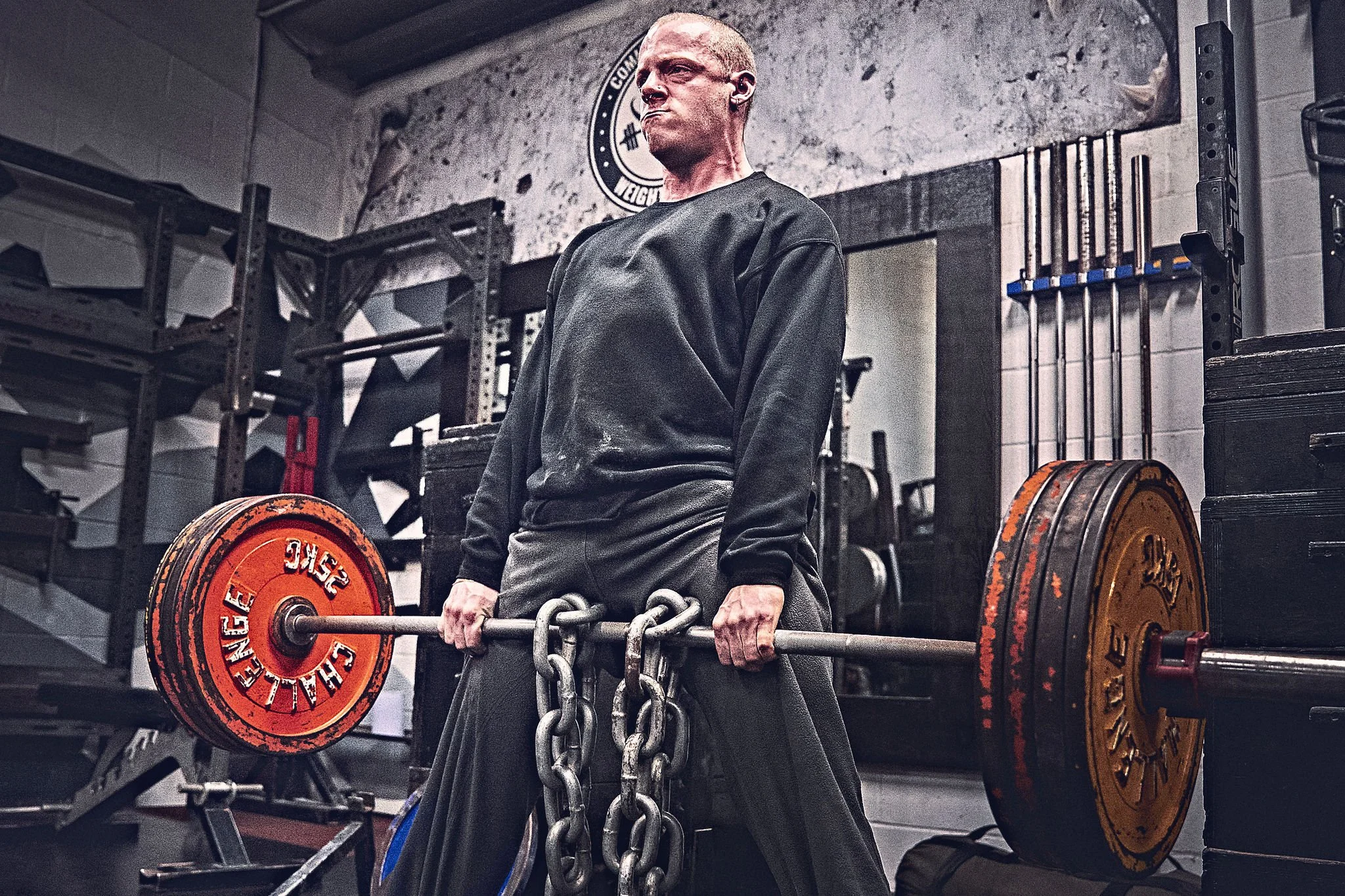 A person with a shaved head and earrings lifting heavy weights in a gym, holding a barbell with weights, surrounded by gym equipment and weight plates. Heavy Deadlifting