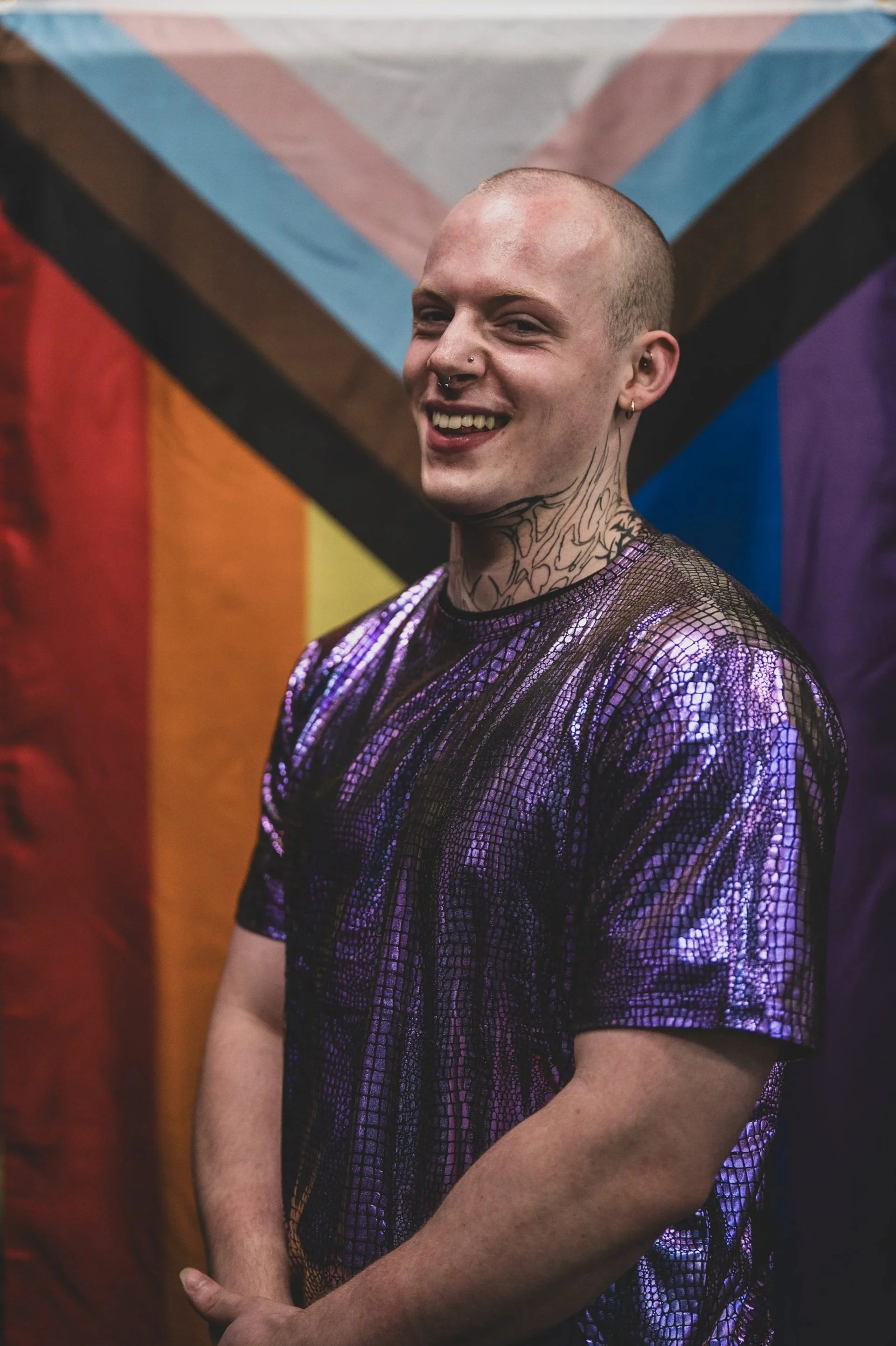 A person with a shaved head, tattoos on the neck, earrings, and nose piercings, wearing a purple and black shimmering snakeskin-patterned top, standing in front of a rainbow Pride flag with pastel and dark colors.