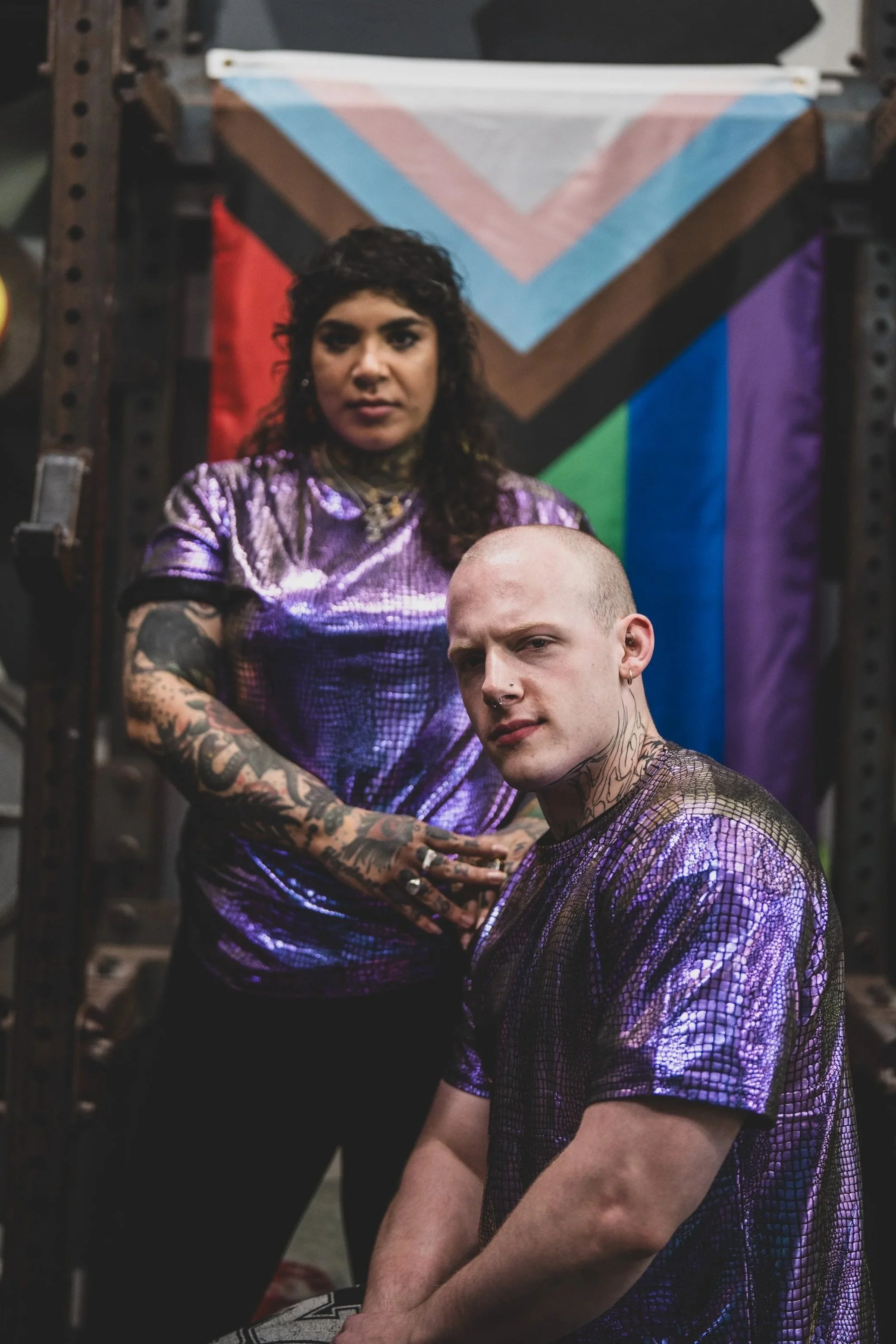 A woman with dark curly hair and tattoos on her arms, wearing a shiny purple top, stands in front of a person with short blonde hair and tattoos, who is sitting cross-legged. Behind them, there is a large colorful flag with a chevron design featuring rainbow colors.