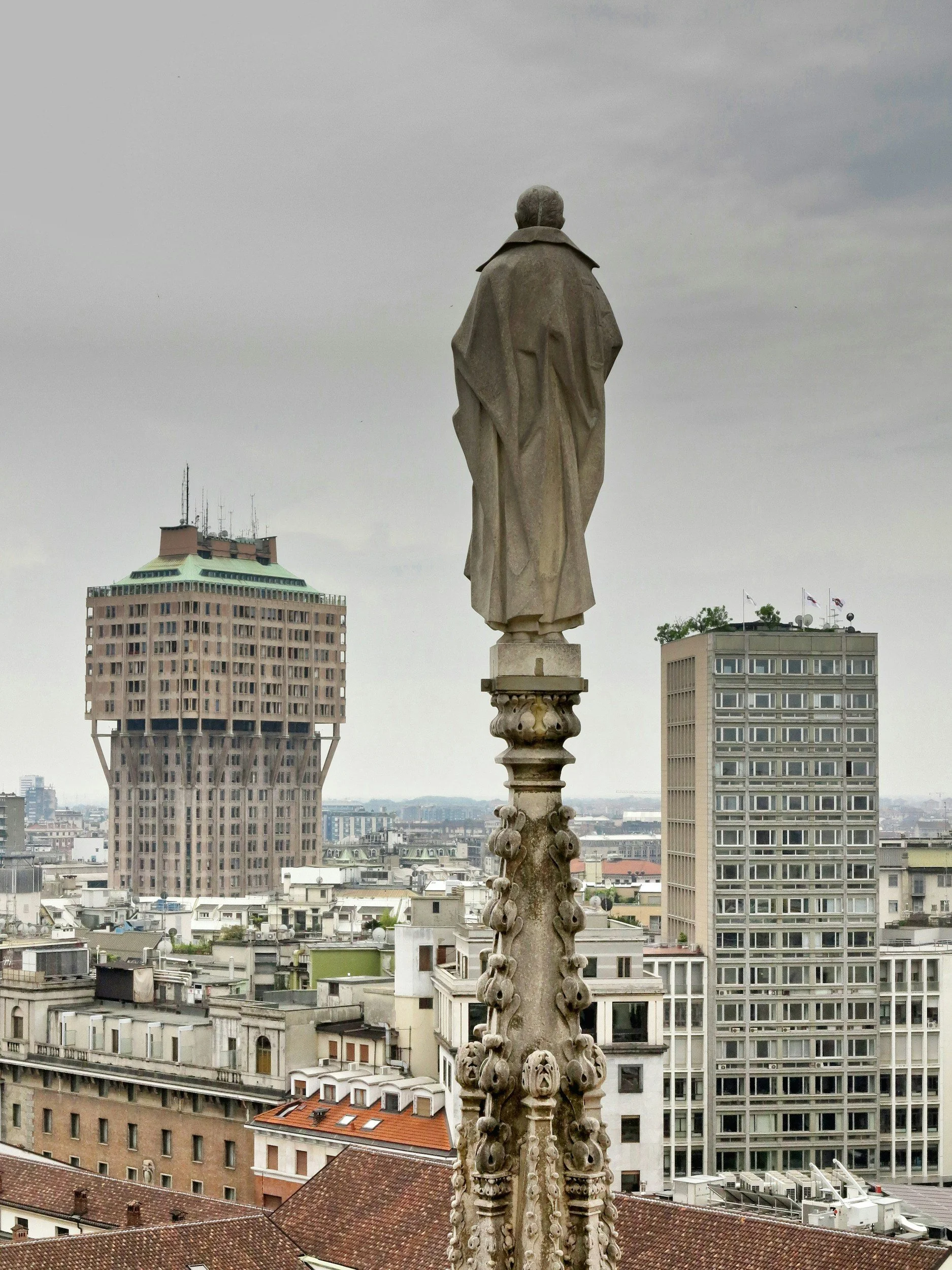 Statua di un personaggio storico situata sulla vetta di una cattedrale, con vista sulla città di Parigi con edifici moderni e storici, cielo coperto.