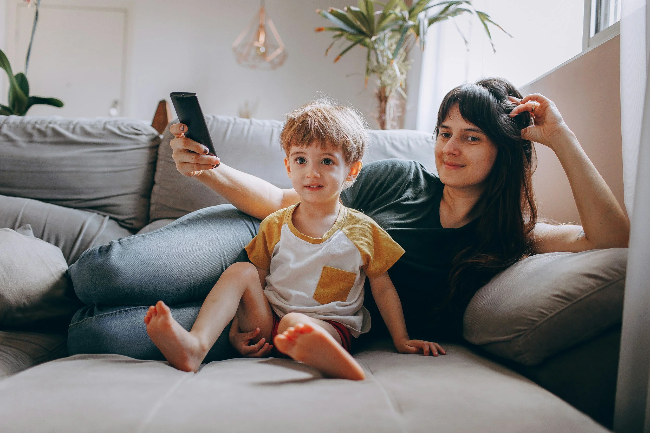 A woman and a young boy with light brown hair, sitting on a gray couch in a bright living room, taking a selfie together. The woman has long dark hair and is smiling while holding her head with one hand. The boy is sitting on her lap, looking at the camera with a neutral expression. The room has large windows, a potted plant, and a modern geometric hanging light fixture.