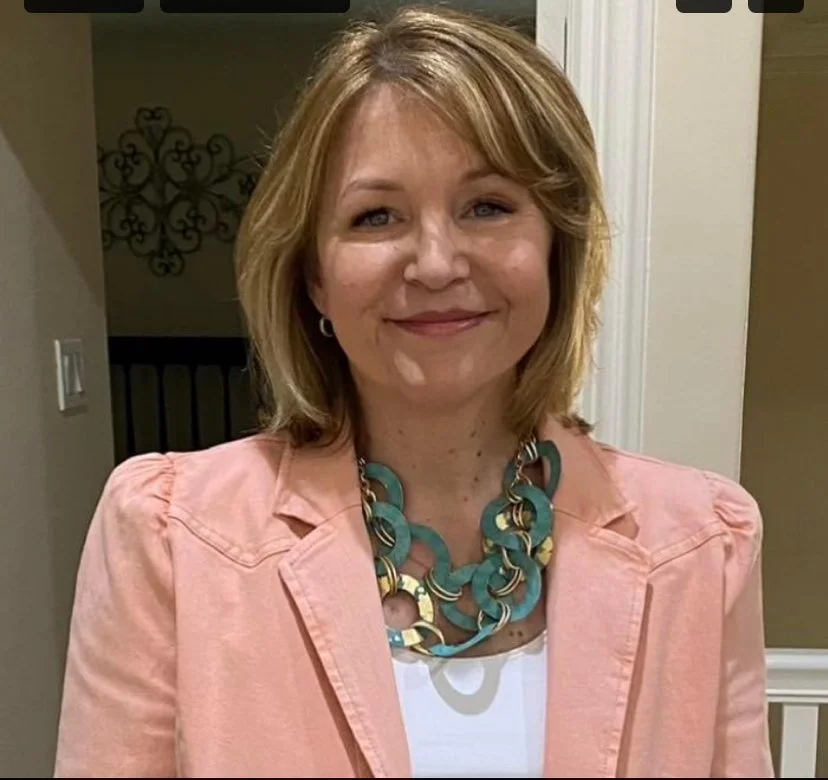 Woman with blonde hair wearing a pink blazer and a large turquoise necklace, smiling indoors.