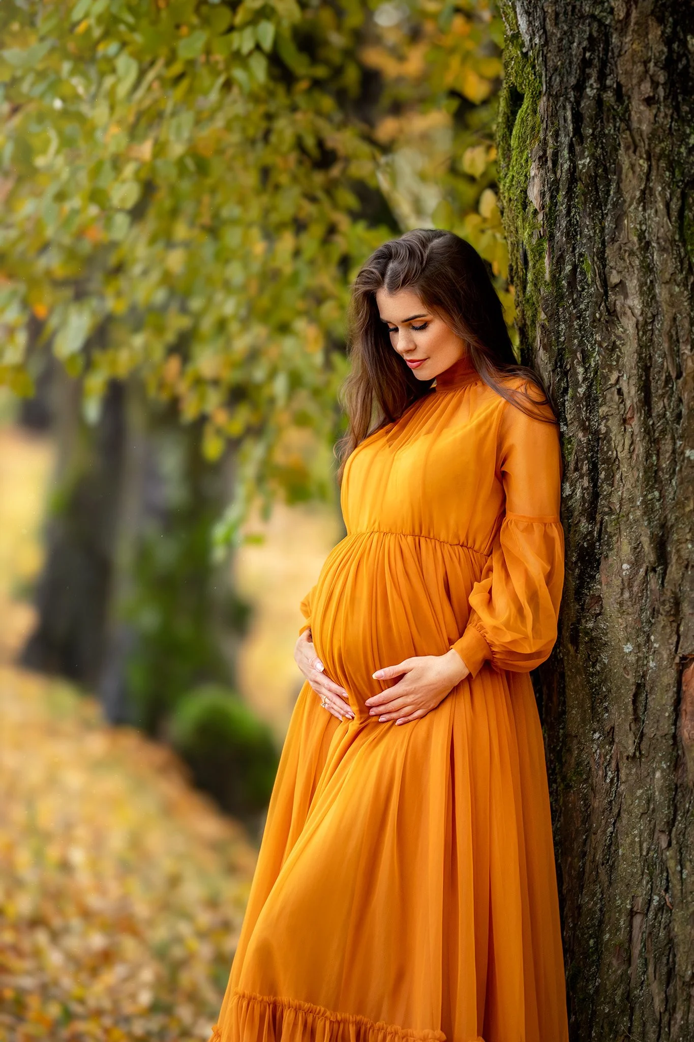 Gravid kvinne i oransje kjole som lener seg mot et tre – gravidfotografering utendørs