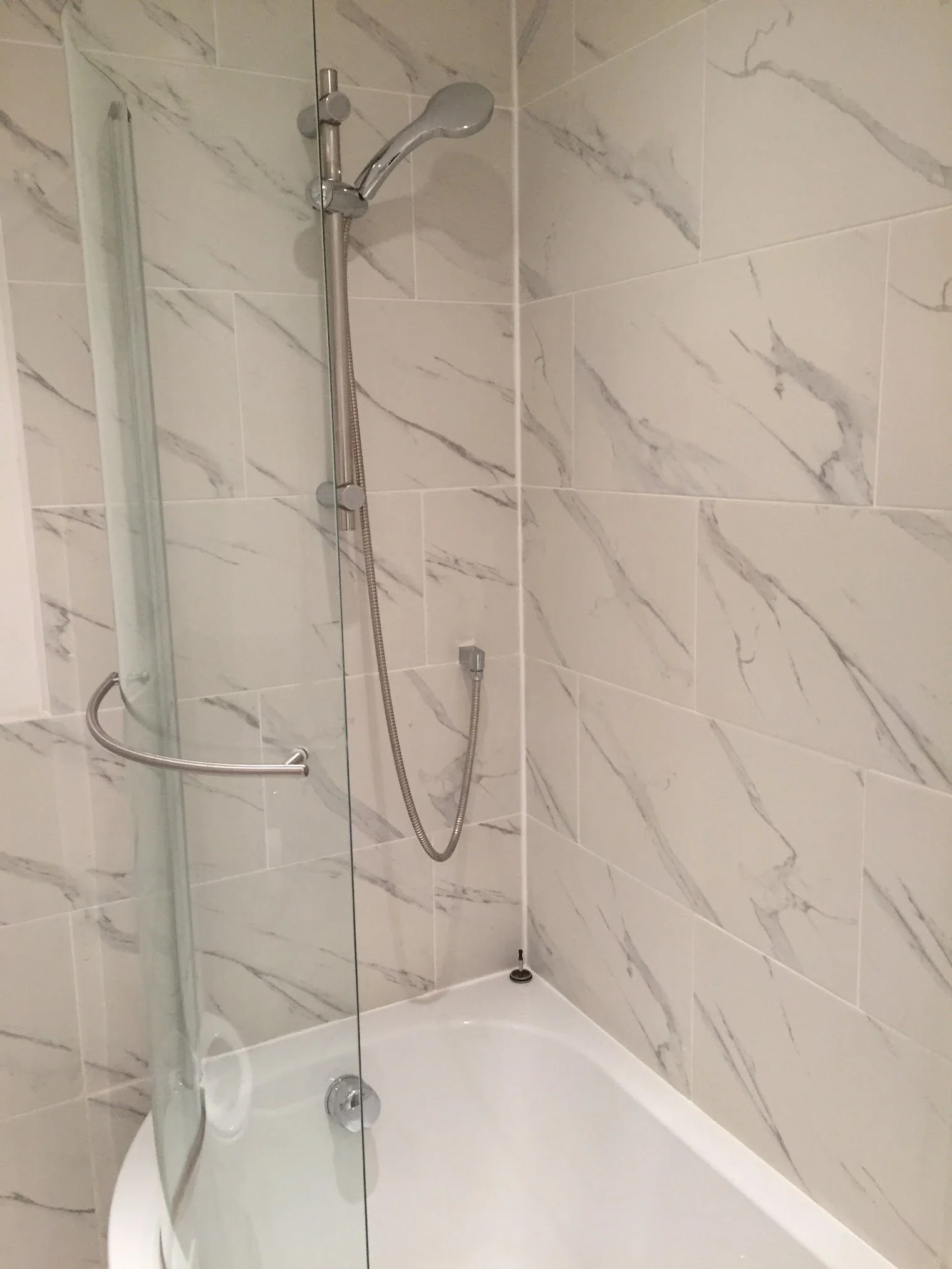 A modern shower with a marble wall, a handheld showerhead, a glass door, and a white bathtub.