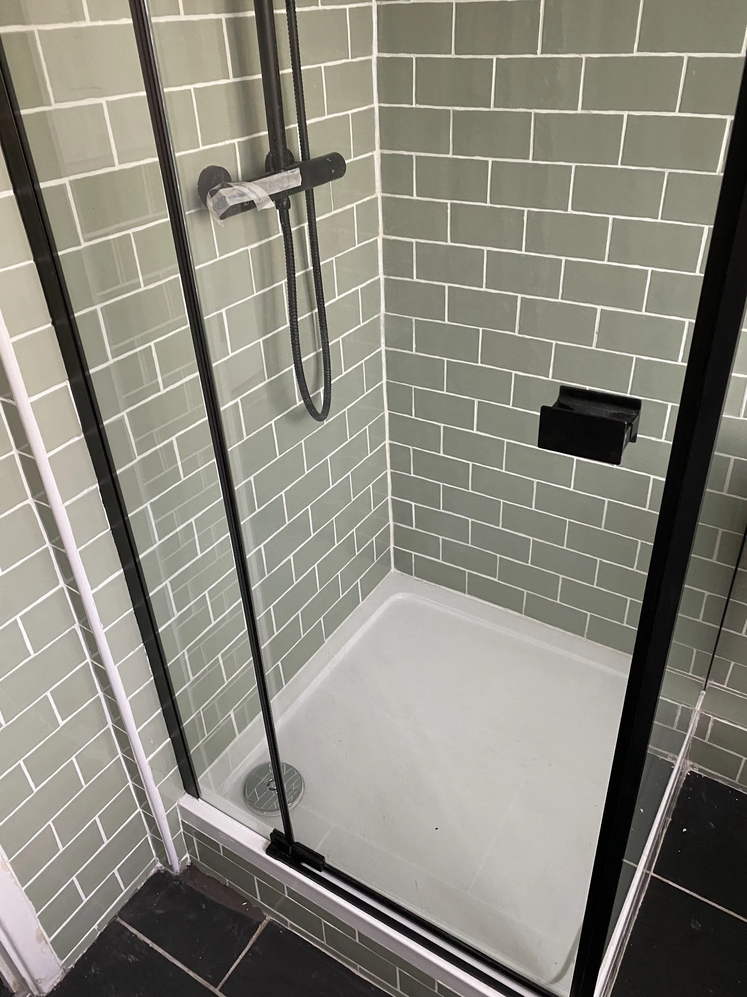 Inside a modern shower with light green subway tiles, glass door, black hardware, fixed black soap dish, and a white shower base with black tile flooring outside.