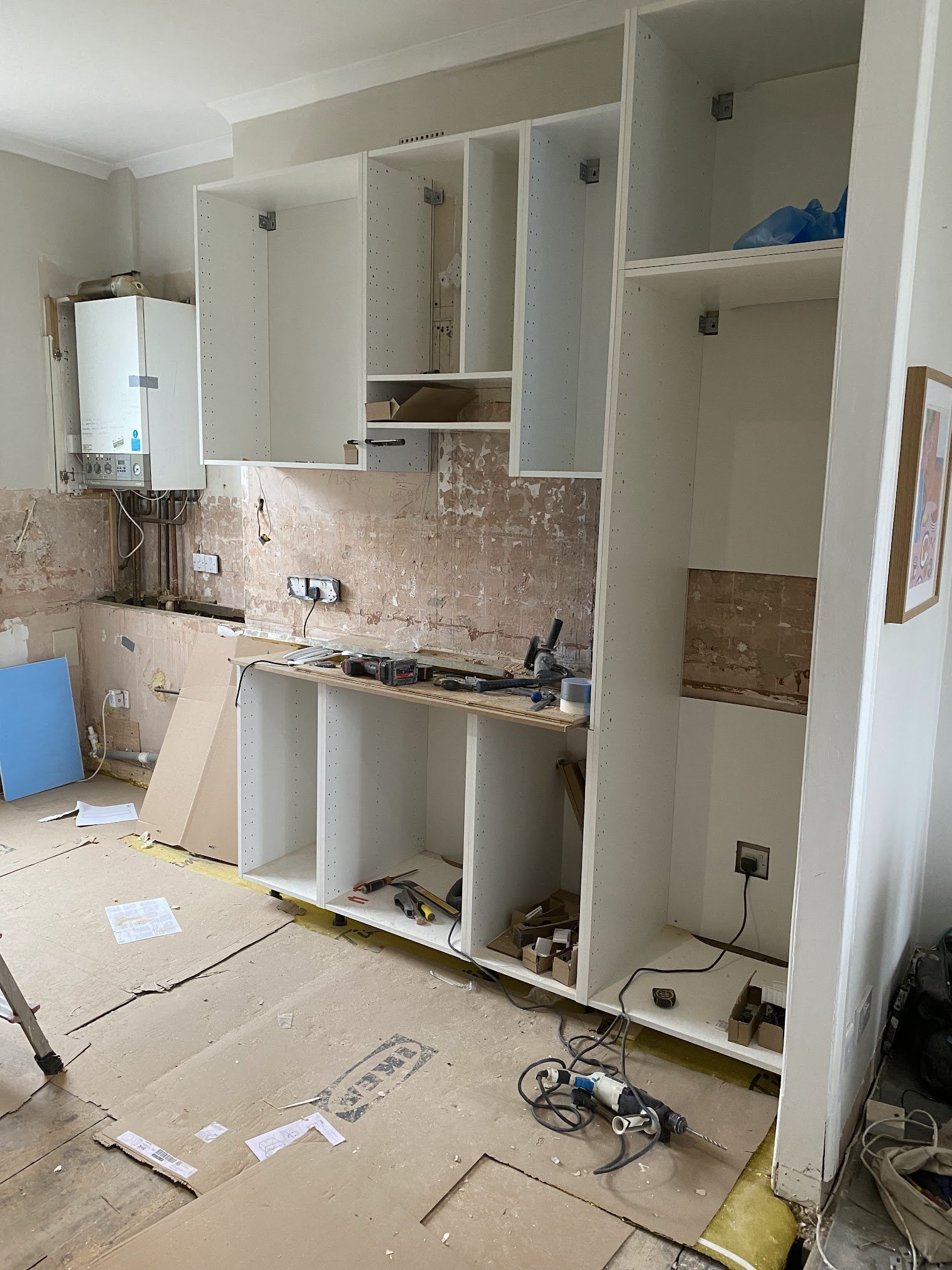 Kitchen cabinets are being installed in a room undergoing renovation. The cabinets are white and partially assembled, with tools and materials scattered on the floor and surface.