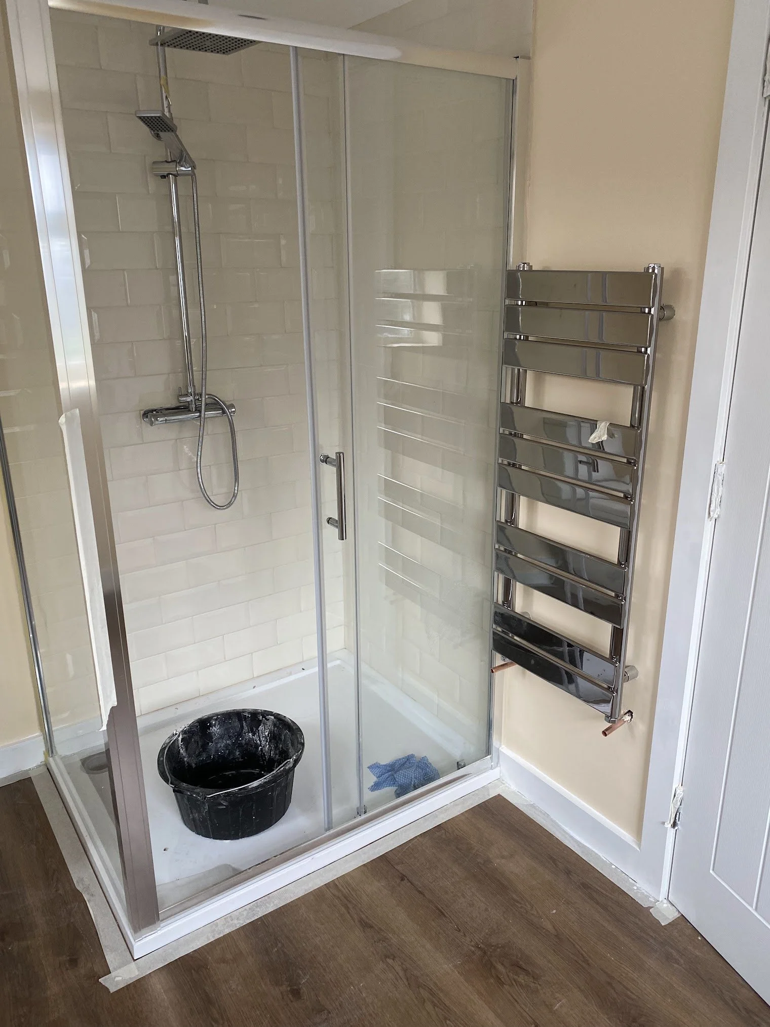 A small bathroom shower with glass door, showerhead, and beige tiles, a black bucket inside, a blue cloth on the shower floor, and a chrome heated towel rail mounted on the wall outside the shower, next to a white cabinet.