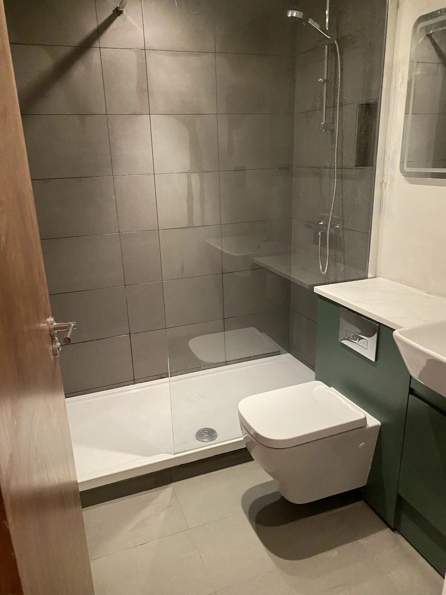 A modern bathroom with beige floor tiles, gray wall tiles, a walk-in shower with a glass door, a white toilet, and a dark green vanity with a sink.